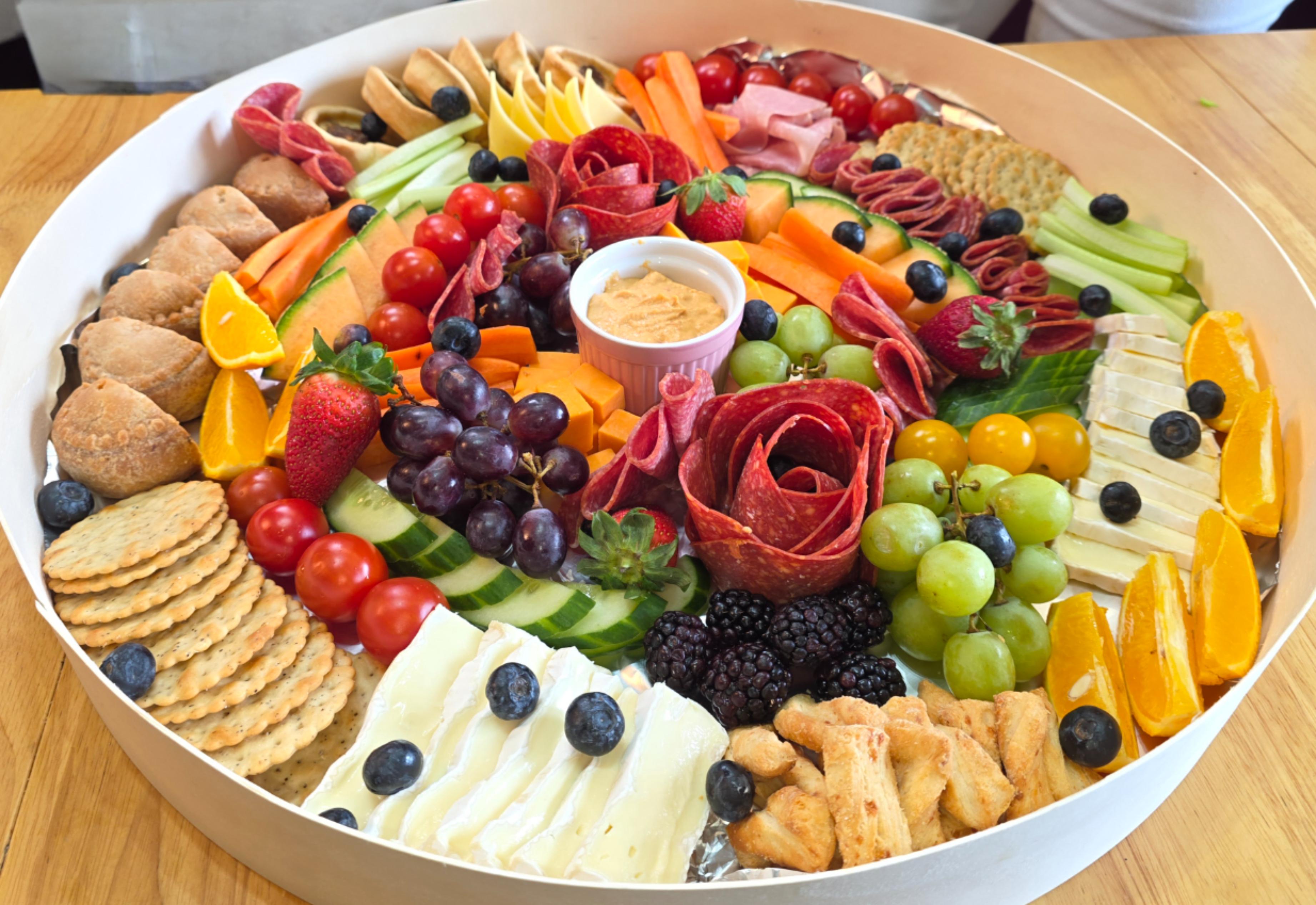 Colorful catering charcuterie tray on a wooden table, with meats, cheeses, crackers, fruit, and dip arranged in a neat circular buffet.