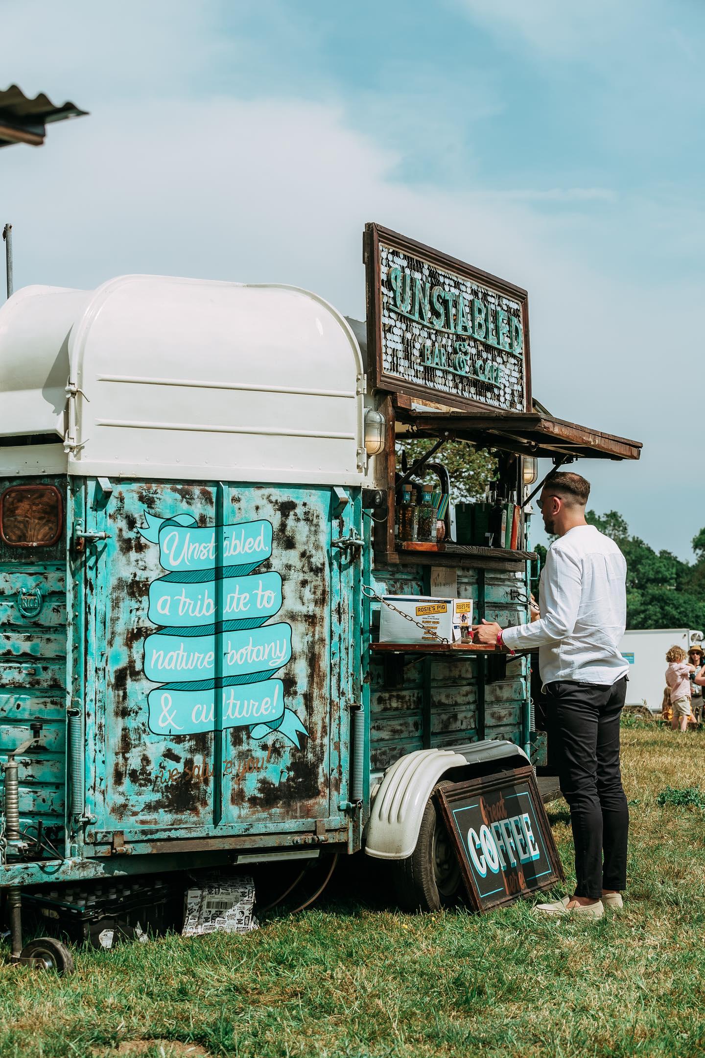A vintage turquoise coffee van with rustic charm, features handwritten signage and a customer ordering in bright daylight on lush green grass.