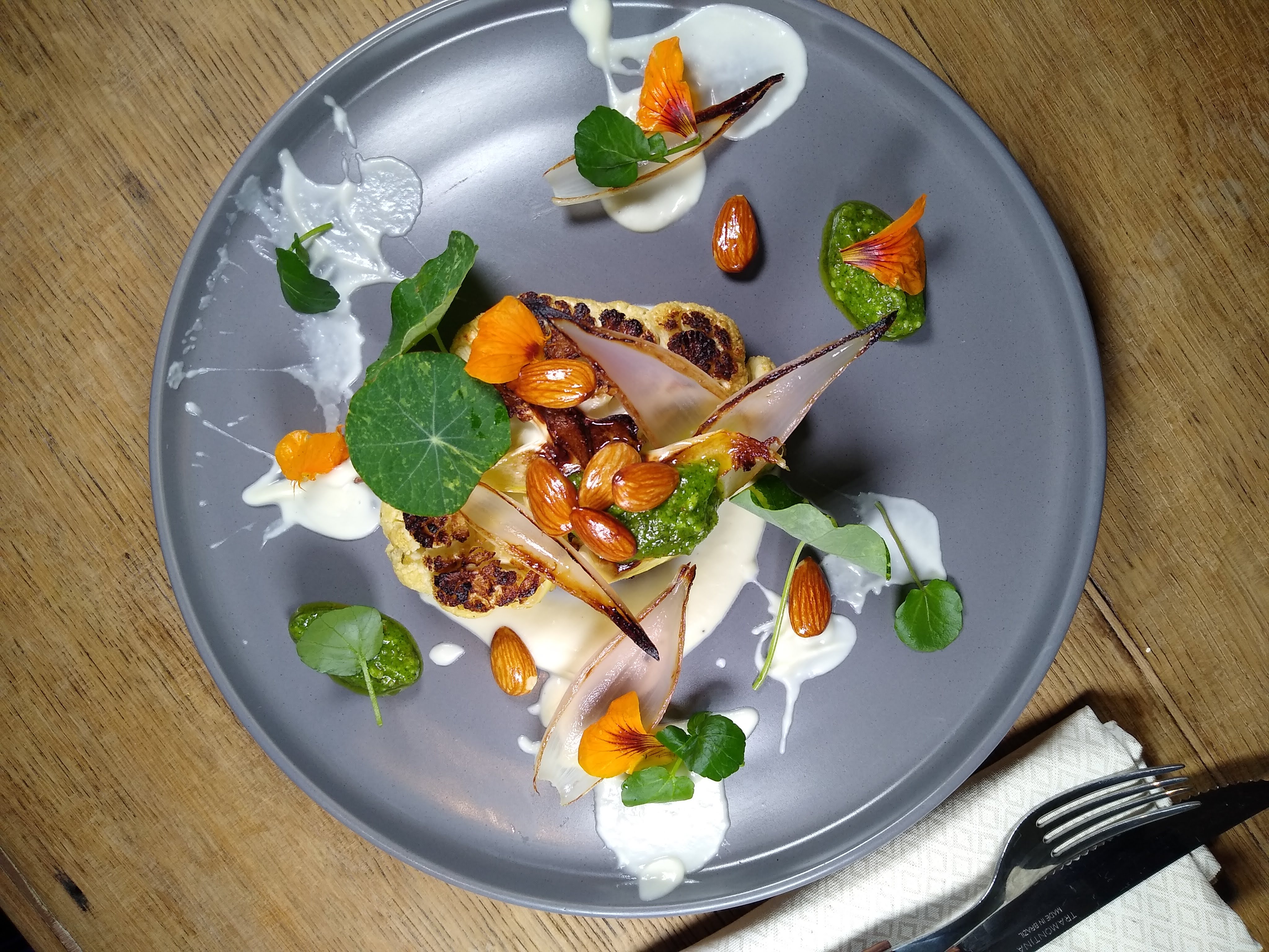 A garnished plate features charred cauliflower, drizzled with creamy sauces, surrounded by vibrant edible flowers and greens on a rustic wooden table.