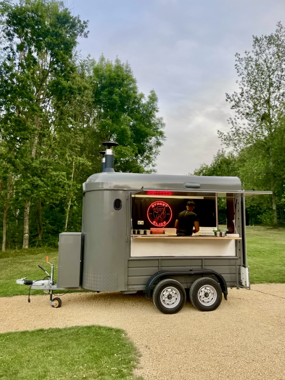 Grey vintage pizza catering trailer with neon Street Slice sign, polished service window, and chef under soft daylight in a park.