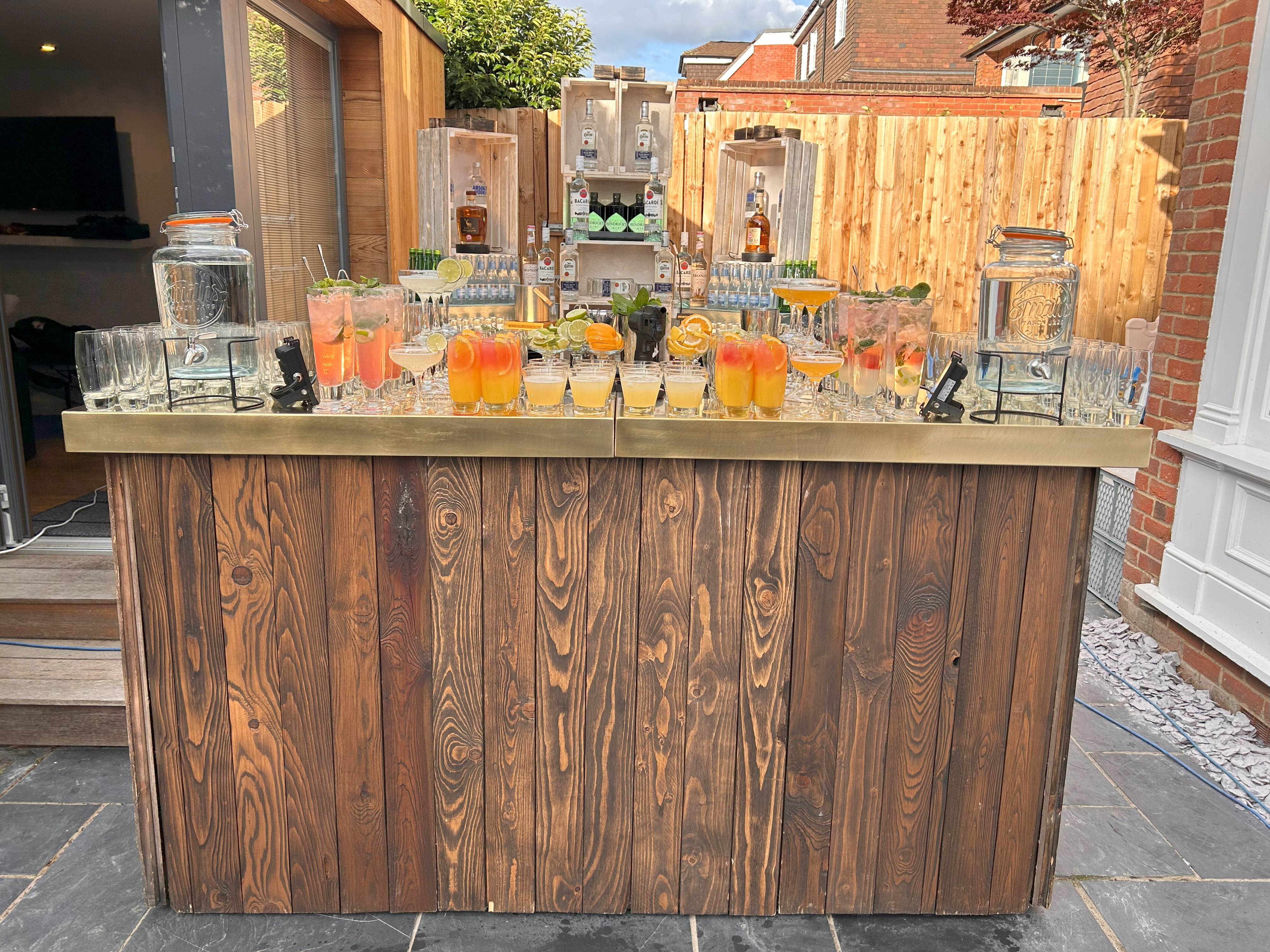 A rustic wooden bar showcases vibrant cocktails in glassware, surrounded by polished accessories, under bright, inviting daylight.