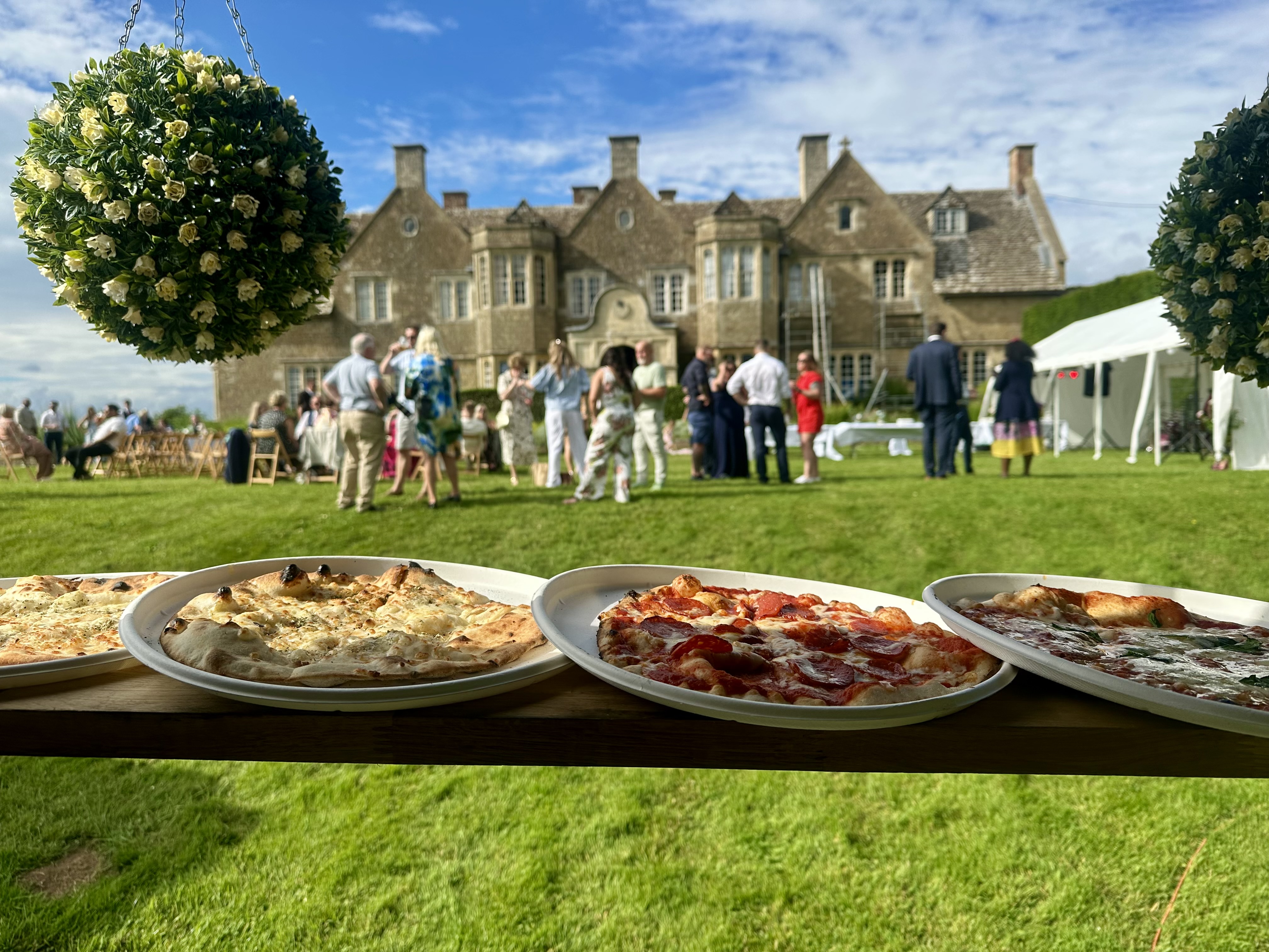 Open-air catering buffet with pizzas on white platters over a wooden table, at a lawn party beside a historic manor under bright daylight.