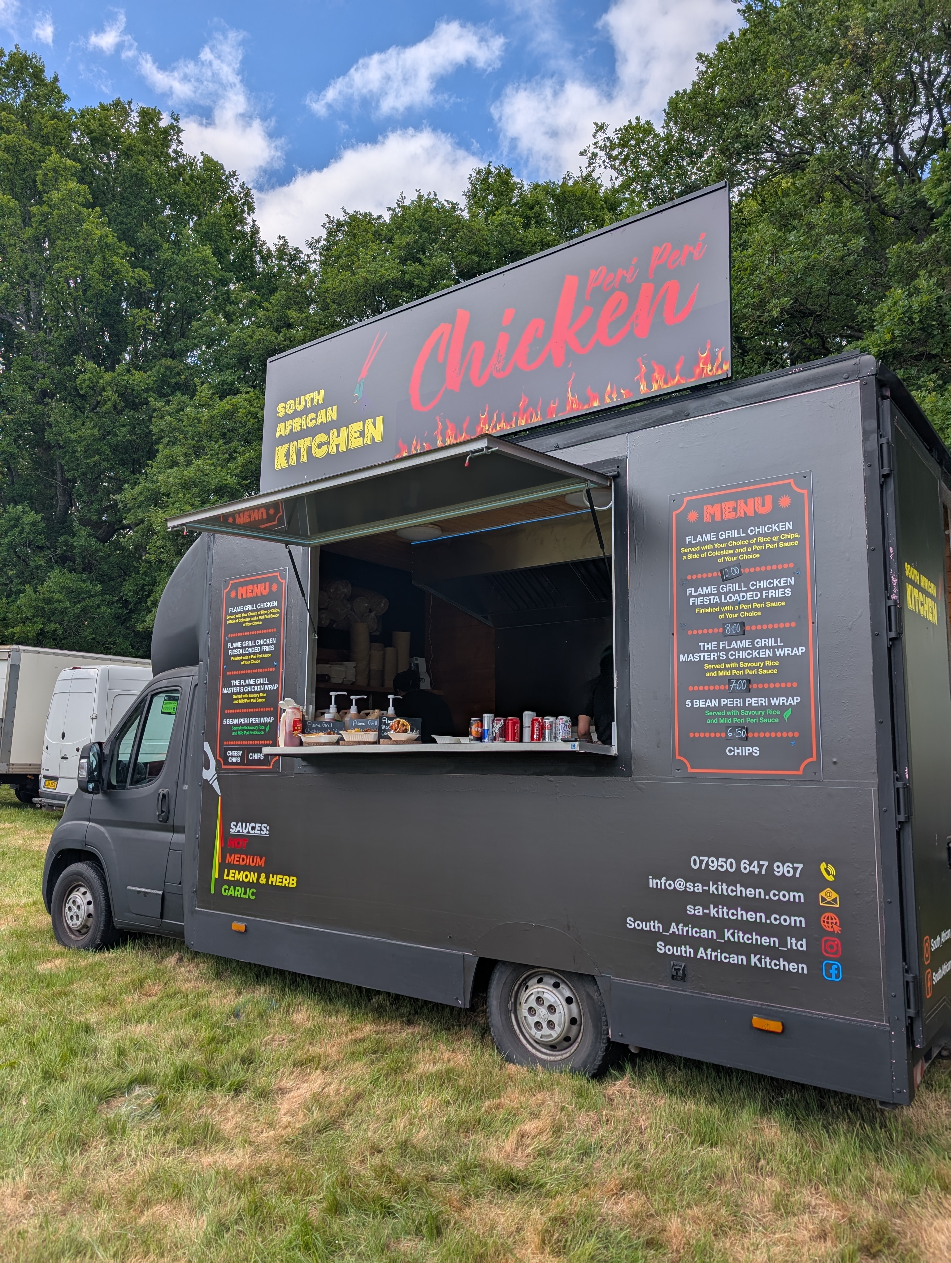 A vibrant food truck boasts a menu featuring flame-grilled peri-peri chicken and loaded wraps, set against lush green trees under a clear sky.