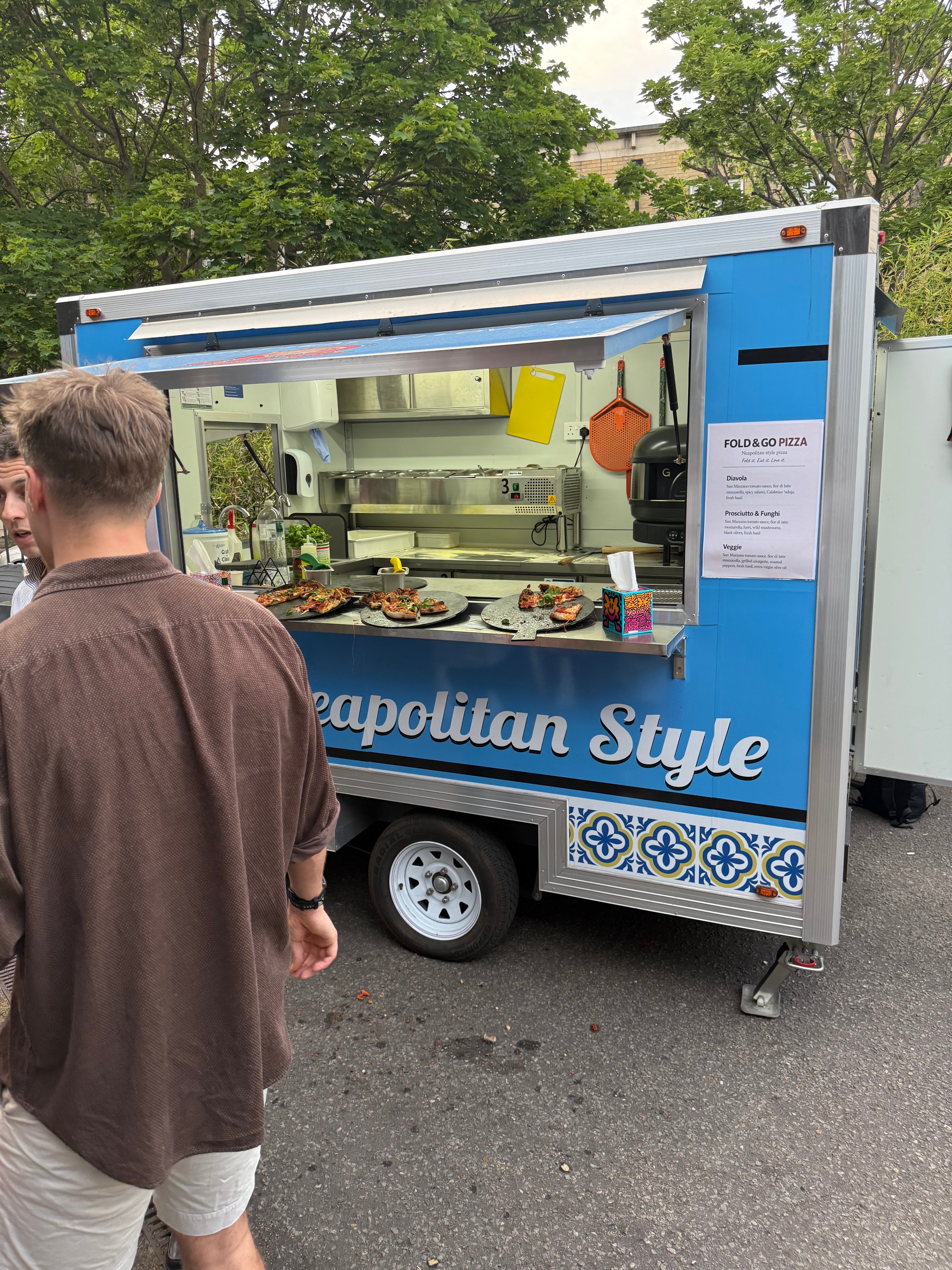 Blue Naples-style pizza truck serving catered slices on slate paddles, with polished stainless gear and bright daylight.