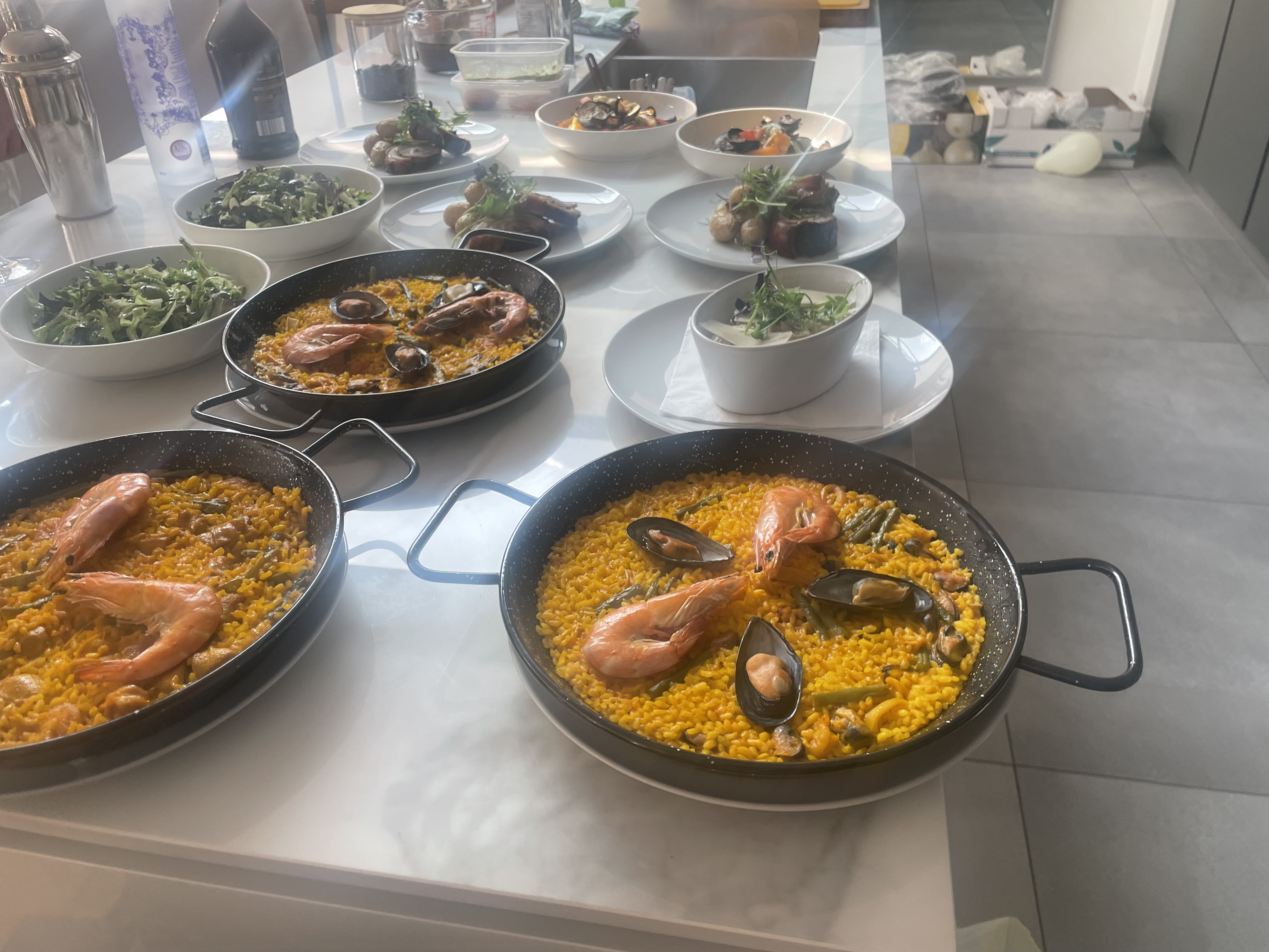 A bright, modern table displays vibrant paella with shrimp and mussels in three shallow pans, surrounded by fresh salads and elegant glassware.