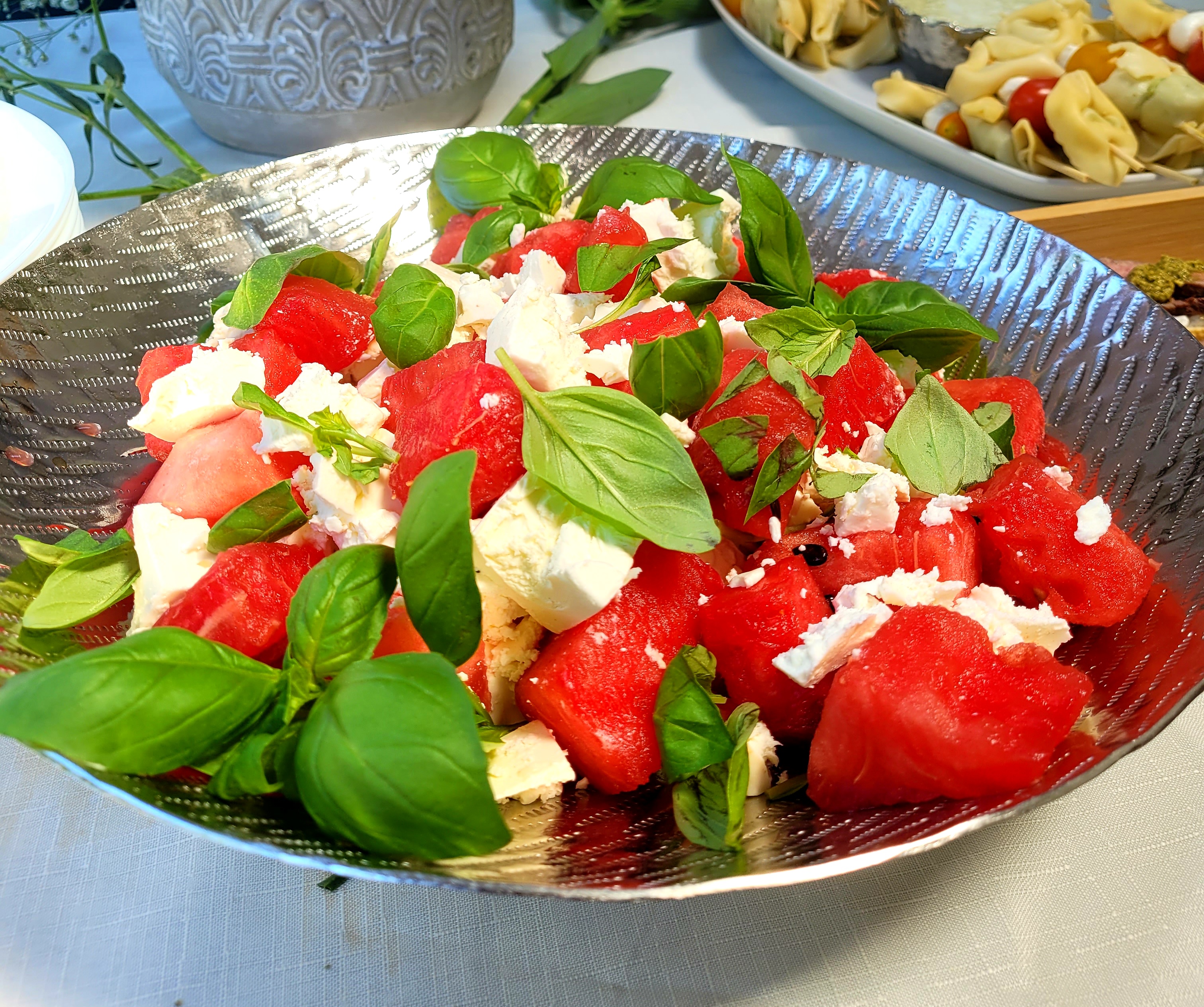 Catering buffet with a hammered silver bowl of watermelon, feta and basil, artfully piled on a white linen table in bright daylight.