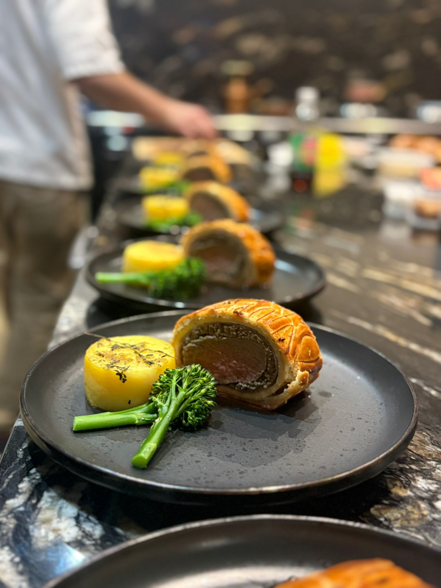 Expertly plated beef Wellington slices, vibrant green broccolini, and yellow potato cakes on sleek black plates, set against a polished marble backdrop.