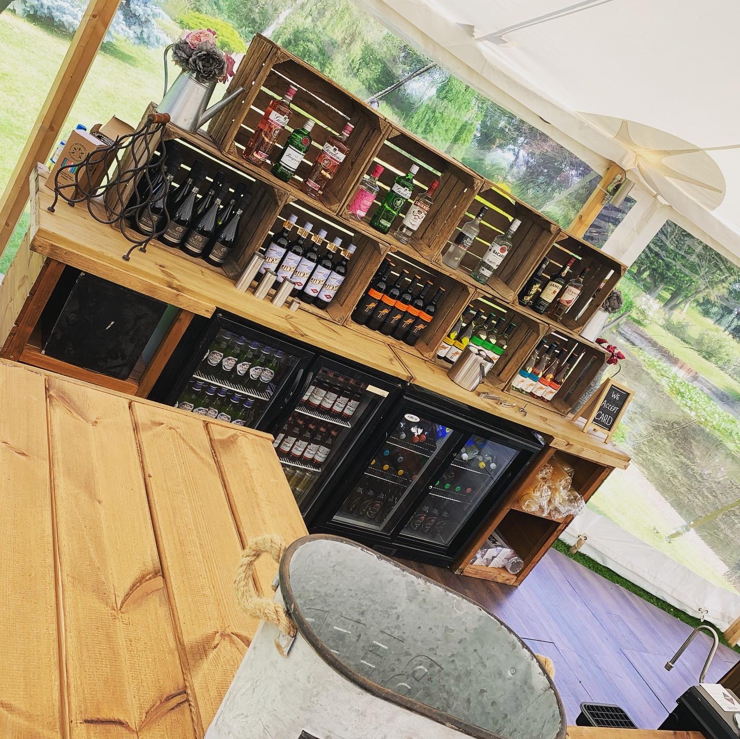 A rustic bar setup features wooden crates brimming with colorful bottles, polished black fridges, and a warm, inviting atmosphere under soft tent lighting.