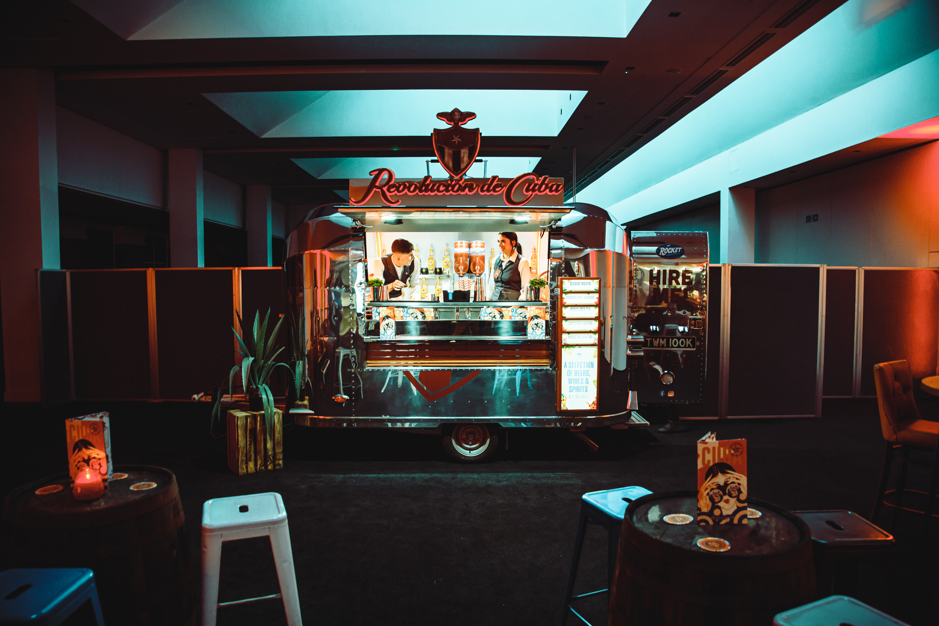 A sleek silver vintage bar truck, "Revolution de Cuba," showcases colorful spirits, with two bartenders amid moody, dim lighting and rustic barrel seating.