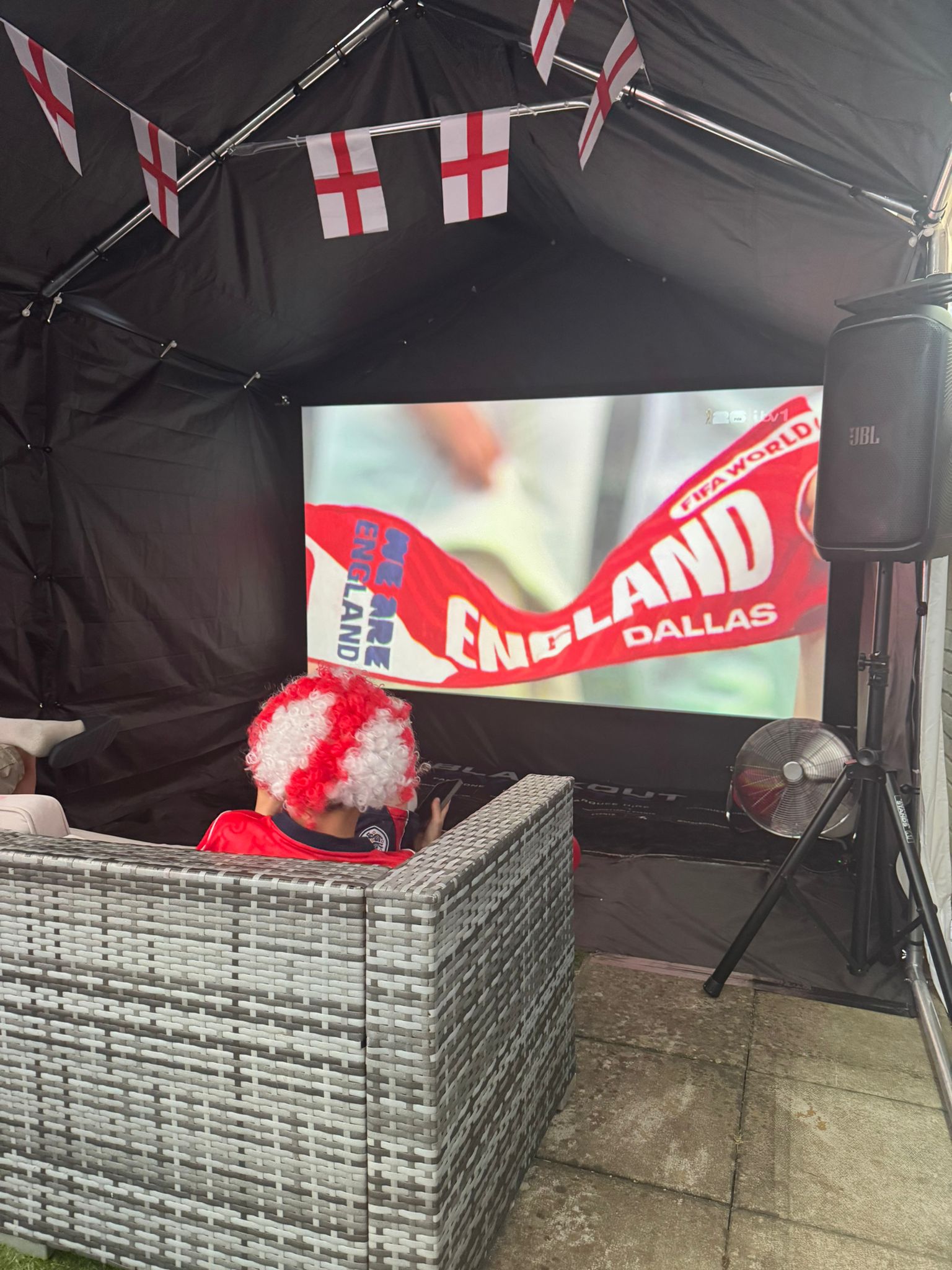 Cozy England football screening tent with black drapes, St George flags, JBL speaker, projector, and wicker sofa, bright daytime event vibe.