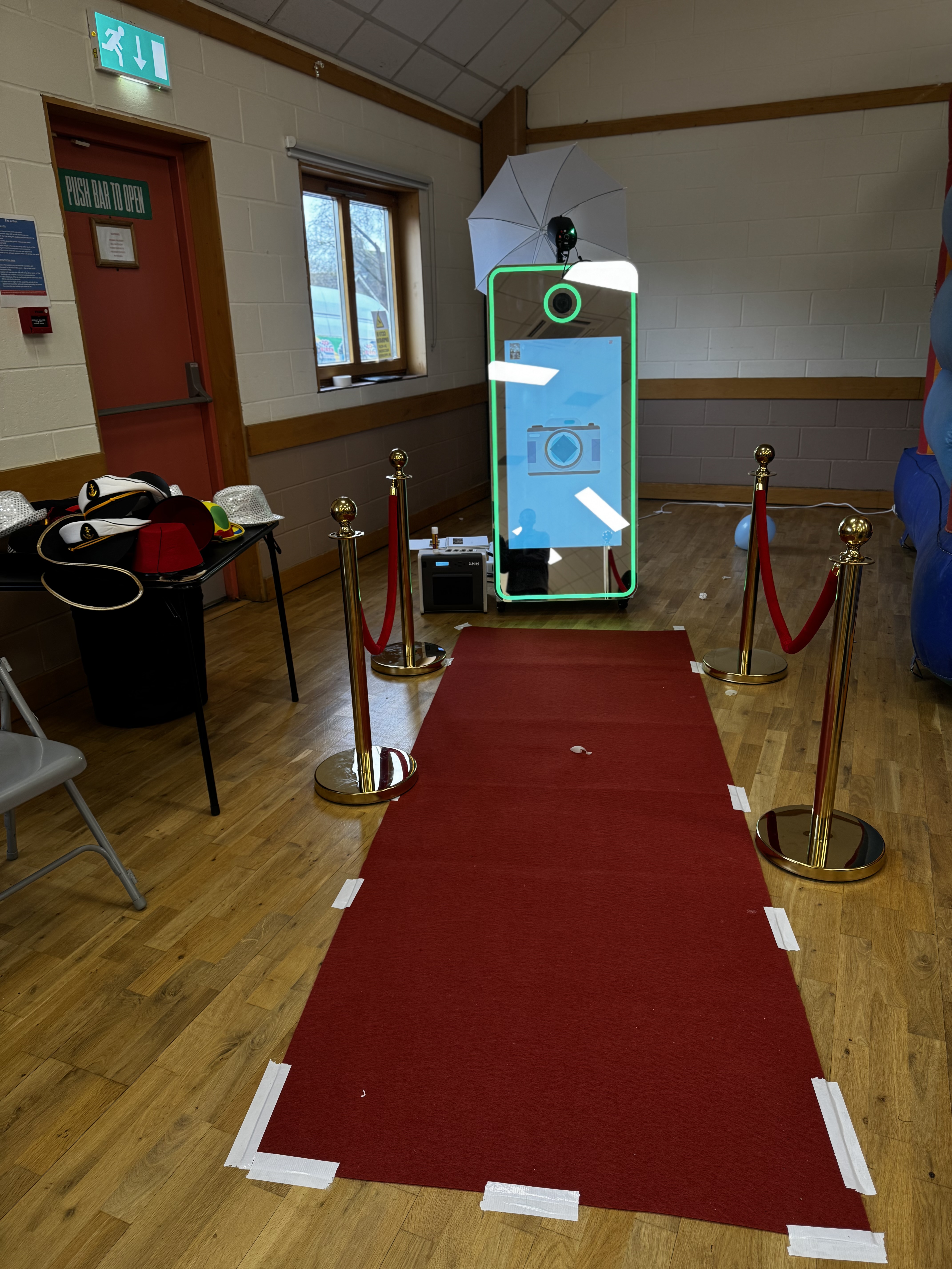 A vibrant photo booth setup features a green-lit mirror, a red carpet flanked by gold stanchions, and playful hats, perfect for events.