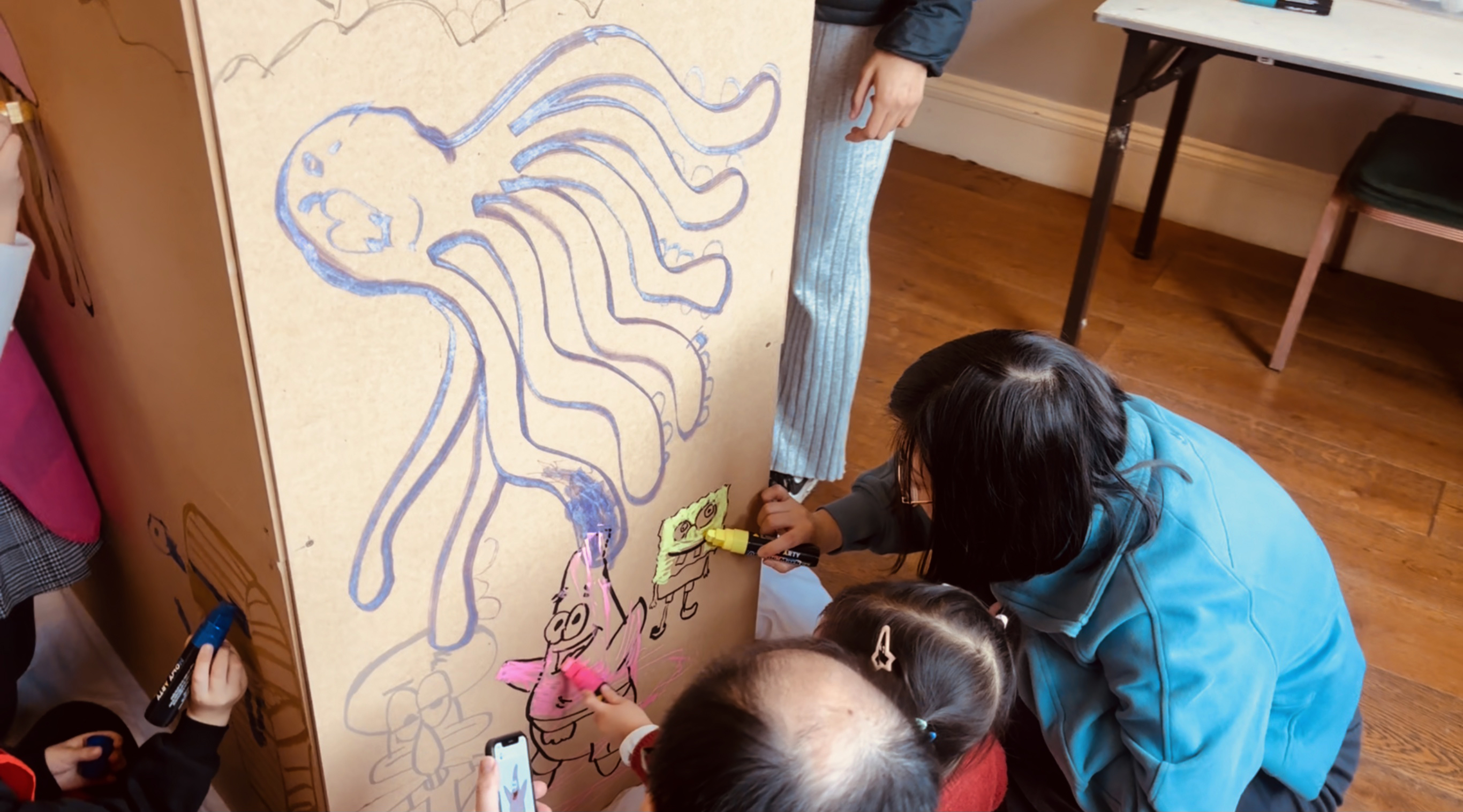 Children enthusiastically color a large cardboard panel with markers, creating playful characters in a bright, inviting room with wooden floors.
