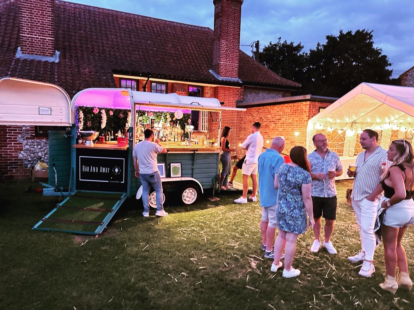 A vintage bar trailer, adorned with twinkling lights, serves cocktails to a lively group enjoying a warm evening in a beautifully manicured garden.
