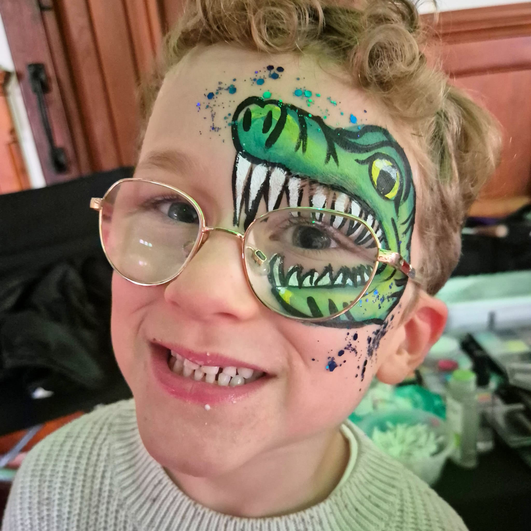 A joyful child with curly hair wears round glasses, showcasing a vibrant green dinosaur face paint adorned with sparkly accents.