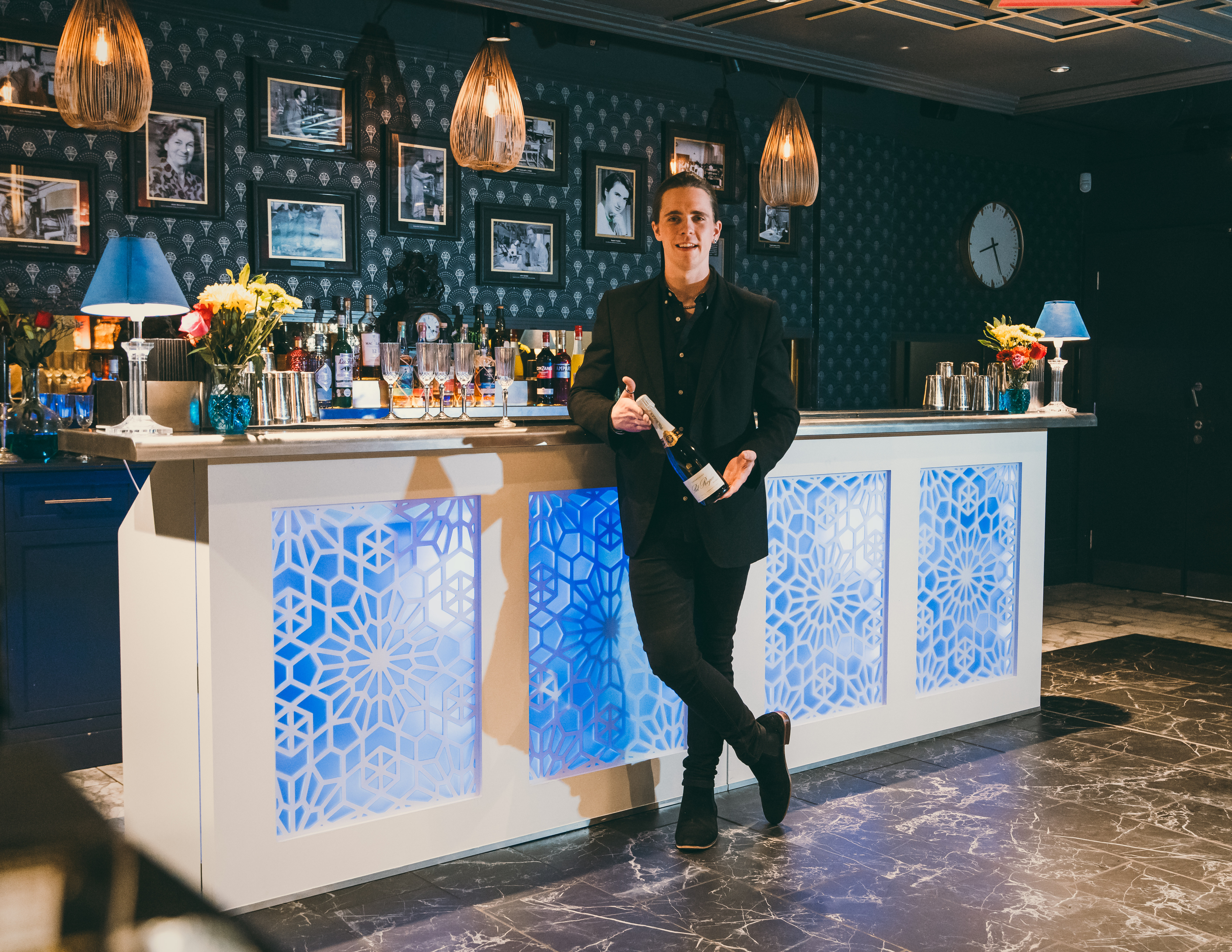 A sleek bar features a lit backdrop, showcasing premium spirits and artful floral arrangements, with warm lighting enhancing the elegant ambiance.