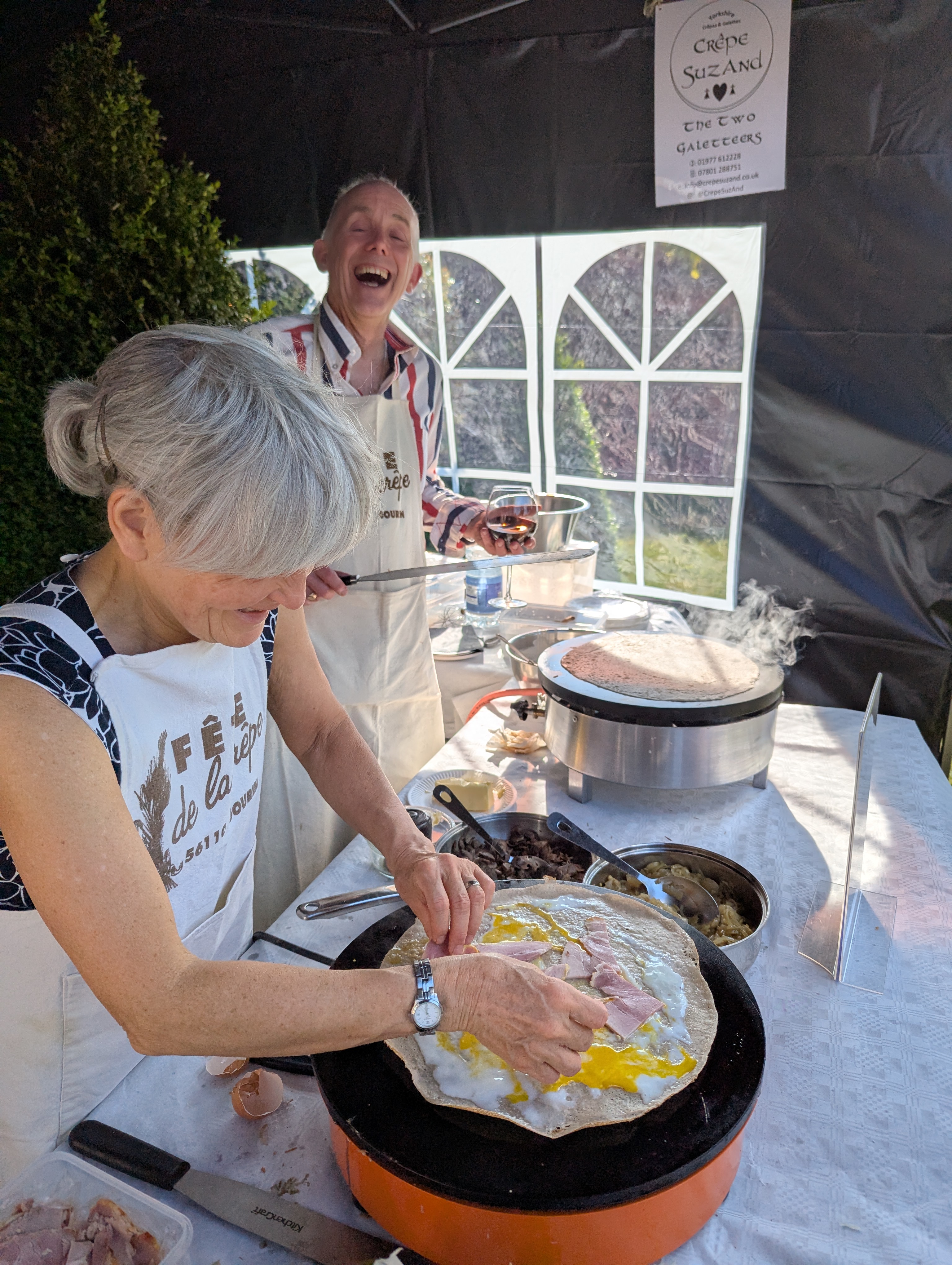 In a sunny outdoor tent, a woman expertly prepares a savory crêpe filled with eggs and ham, while a smiling man holds a glass of wine, radiating warmth.