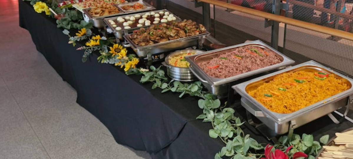 Catering buffet in stainless chafing pans, with rice, meat and dumplings, lined on a black-draped table with floral garland under bright indoor light.