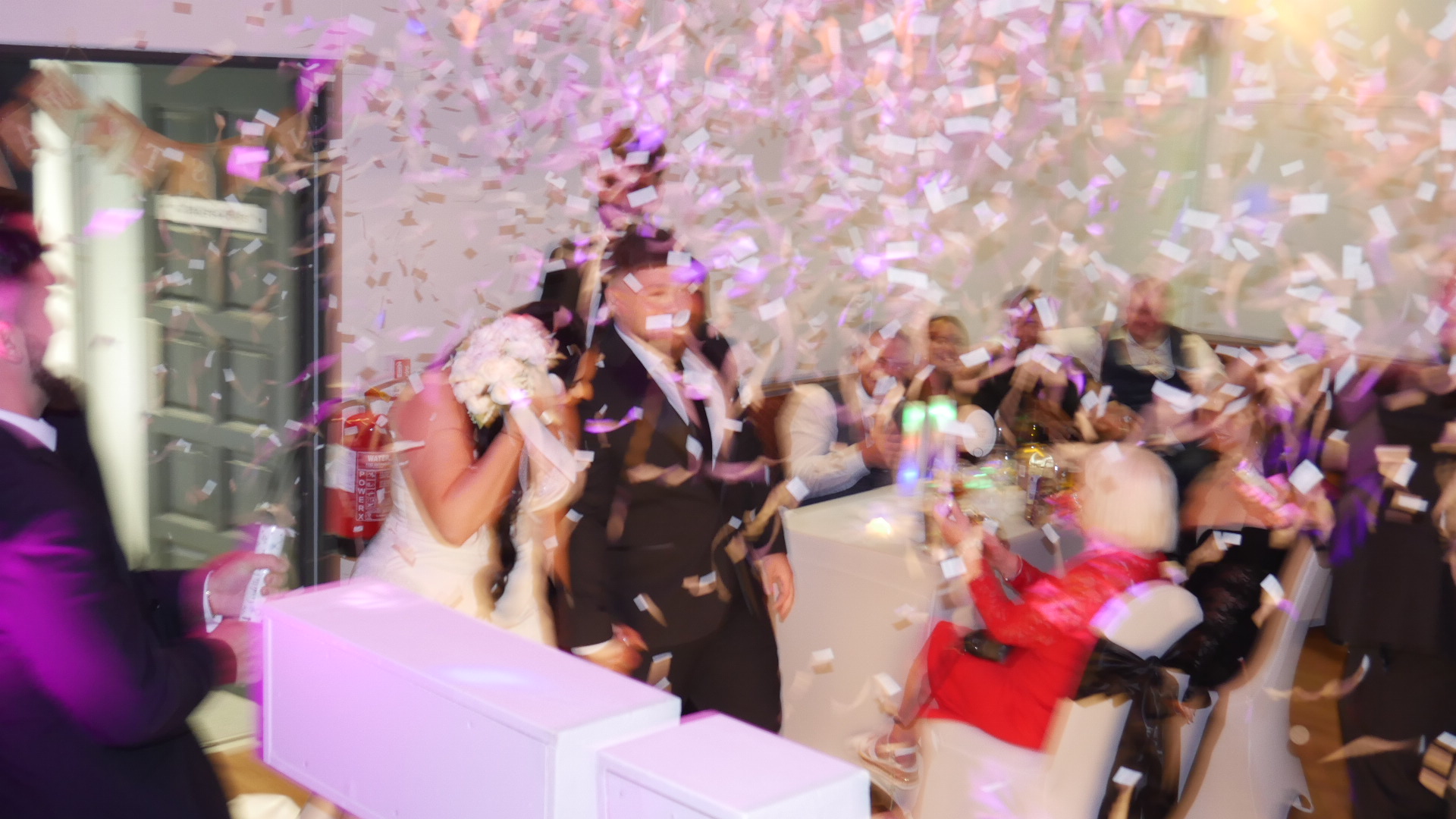 Confetti-filled wedding reception with bride and groom amid seated guests, white linens, polished glassware, and warm celebration lighting.
