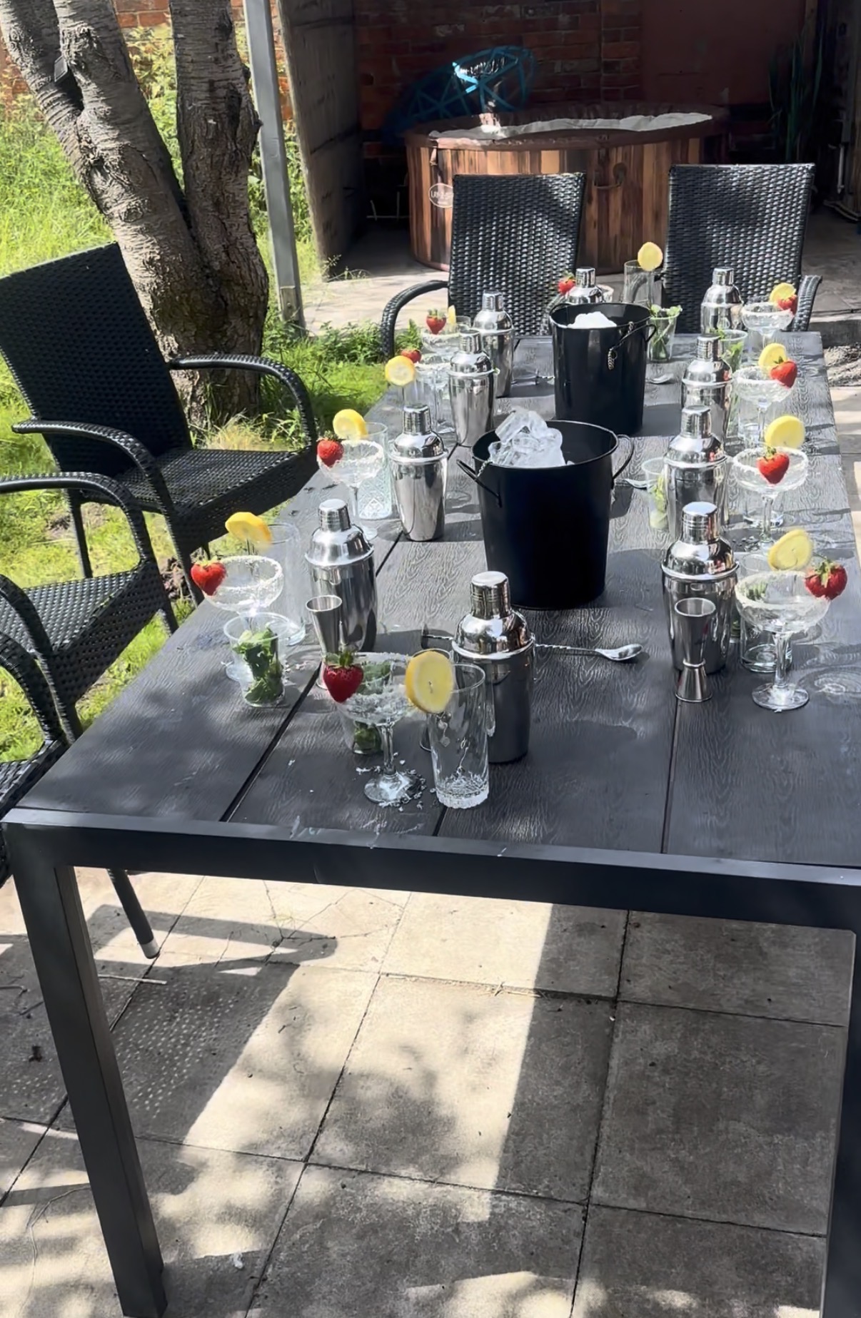 Sunlit outdoor catering setup with black wicker chairs, dark wood table, silver cocktail shakers, ice buckets, and lemon-strawberry garnishes.