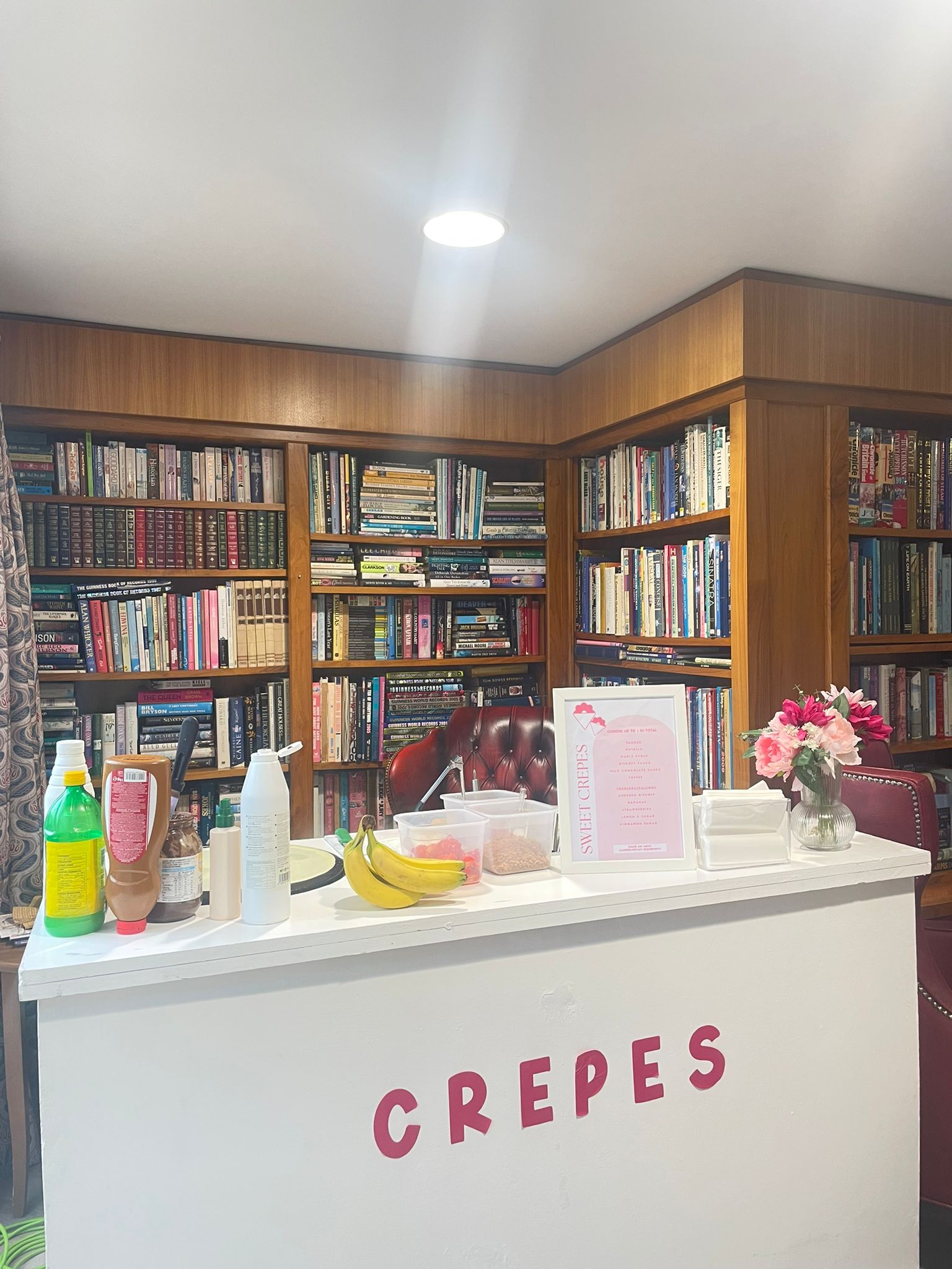A cozy crepe stand features vibrant bananas, chocolate syrup, and neatly arranged sauces in containers against a backdrop of wooden shelves filled with books, all under warm lighting.