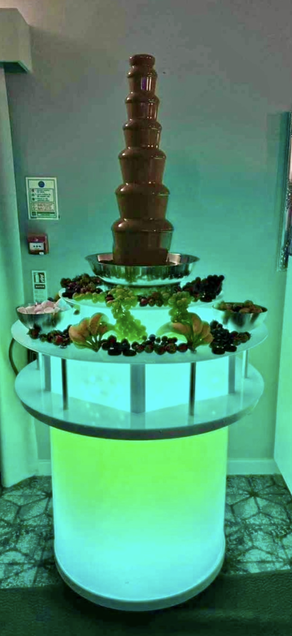 A sleek chocolate fountain stands atop a glowing cylindrical base, surrounded by fresh strawberries, grapes, and marshmallows, creating an inviting dessert display.