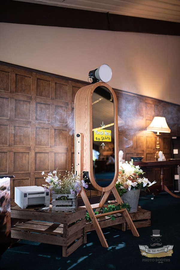 A rustic-style photo booth features a large mirror with a bright touchscreen and white floral arrangements, creating a charming, inviting atmosphere.