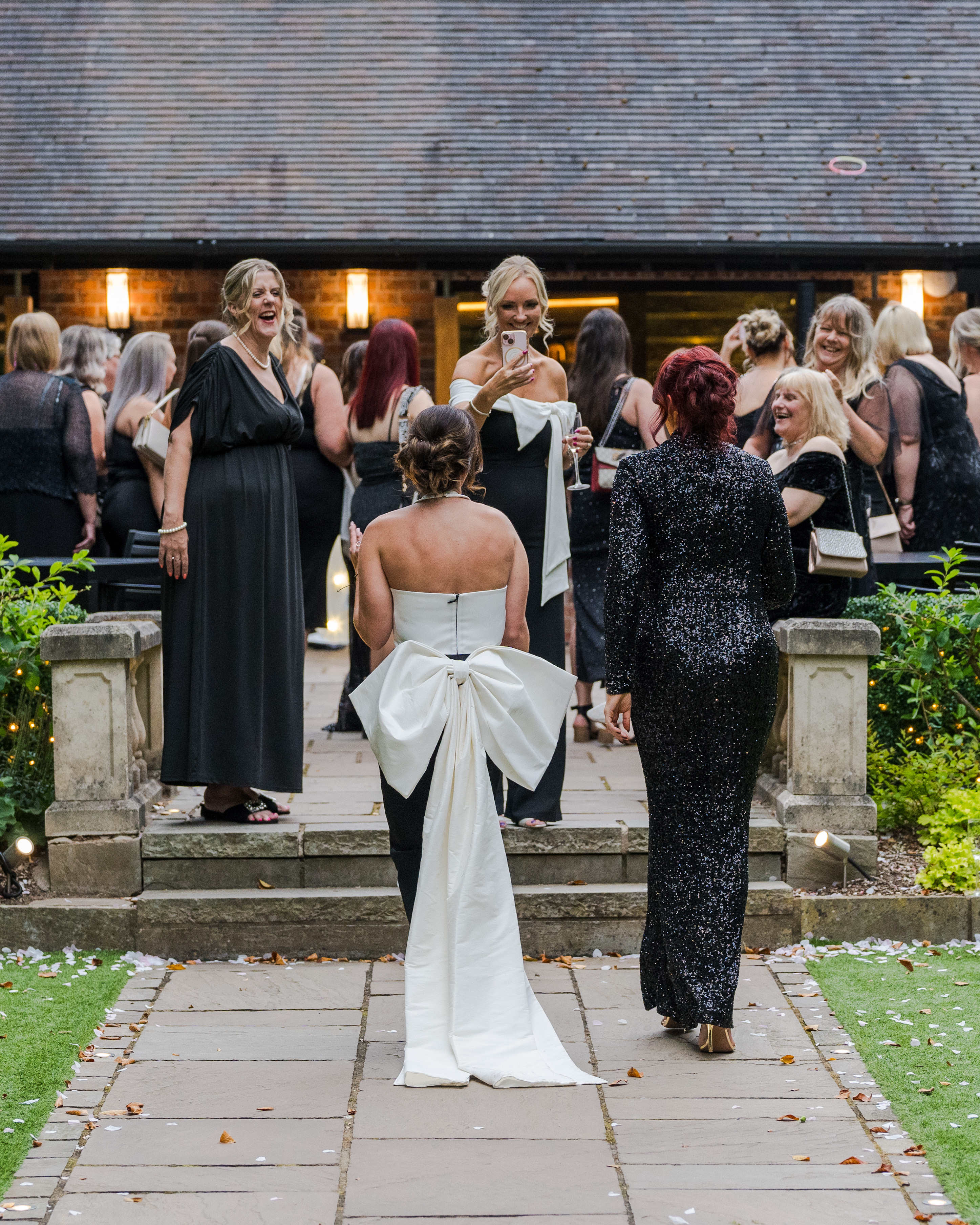 A glamorous outdoor event features elegantly dressed women in black and silver gowns, laughing and mingling under warm evening lights.