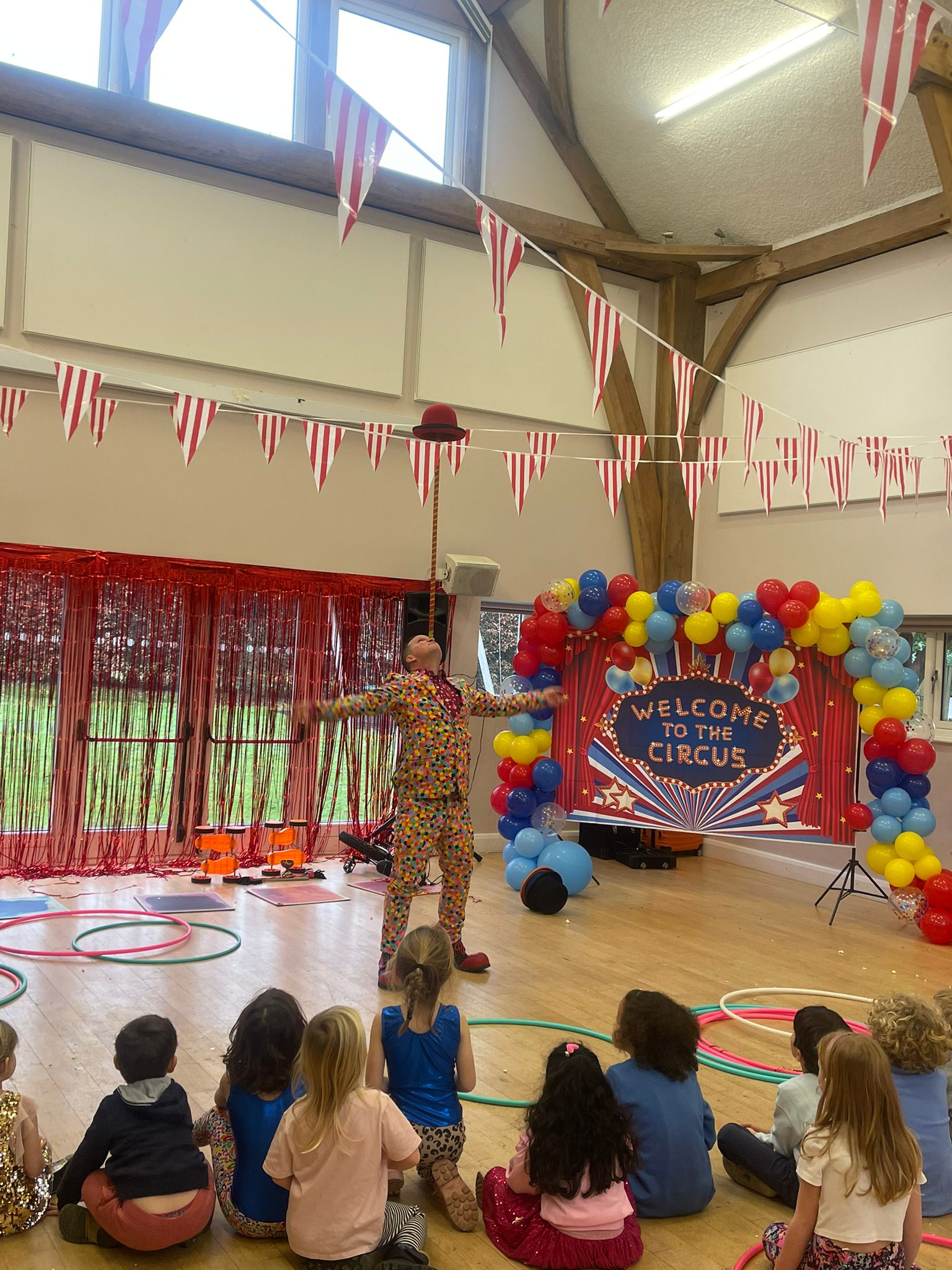 Colorful circus party performance in a bright hall, with balloon arch, striped bunting, hoops, and seated children watching the entertainer.