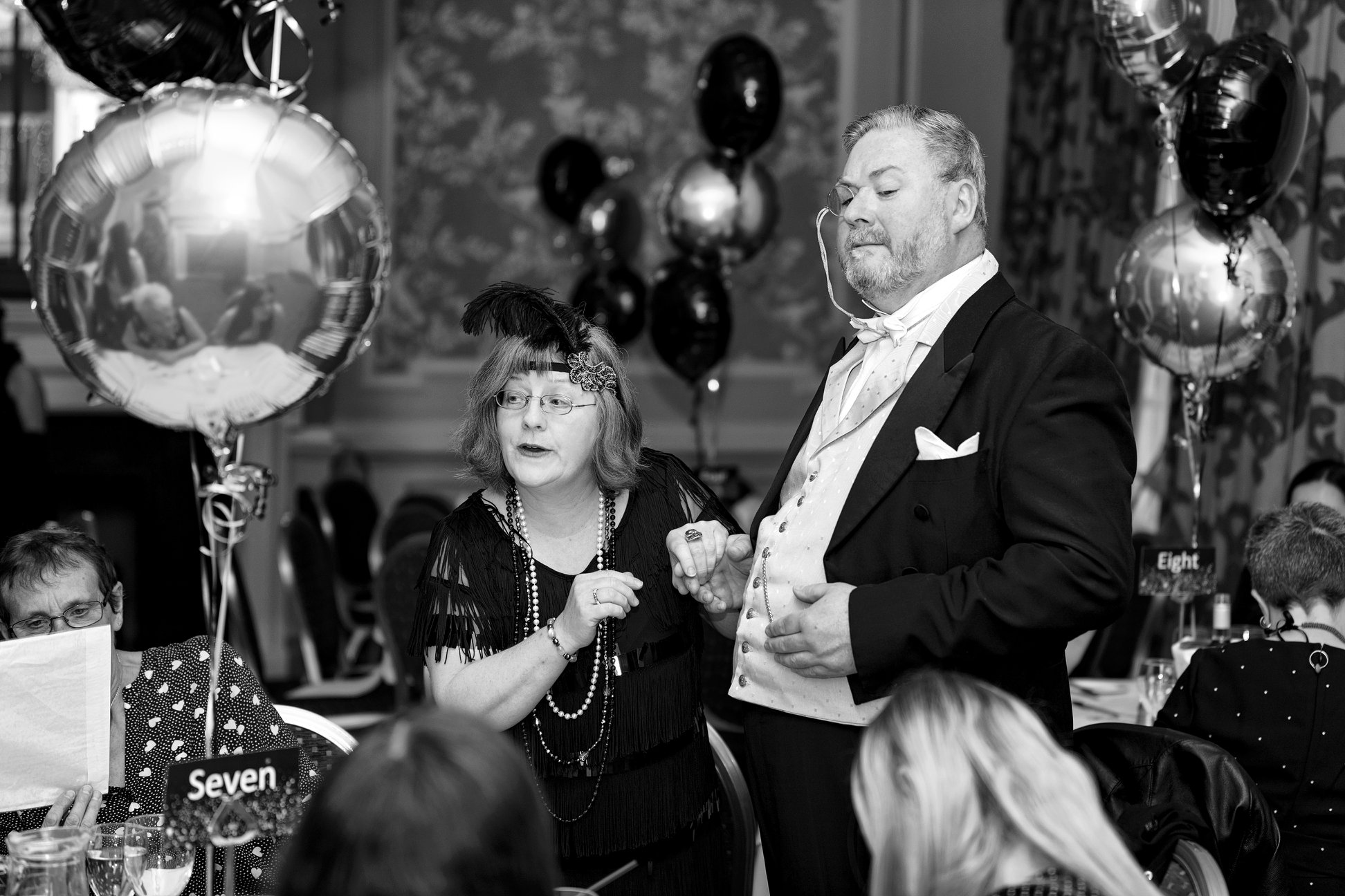 Two elegantly dressed individuals engage in conversation amid an upscale banquet setting, adorned with black and silver balloons and guests.