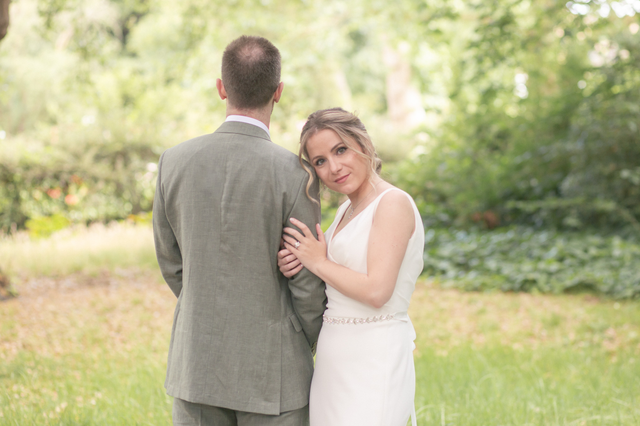 Bride in white hugs groom in sage suit outdoors, soft daylight and leafy garden backdrop creating an elegant wedding portrait vibe.