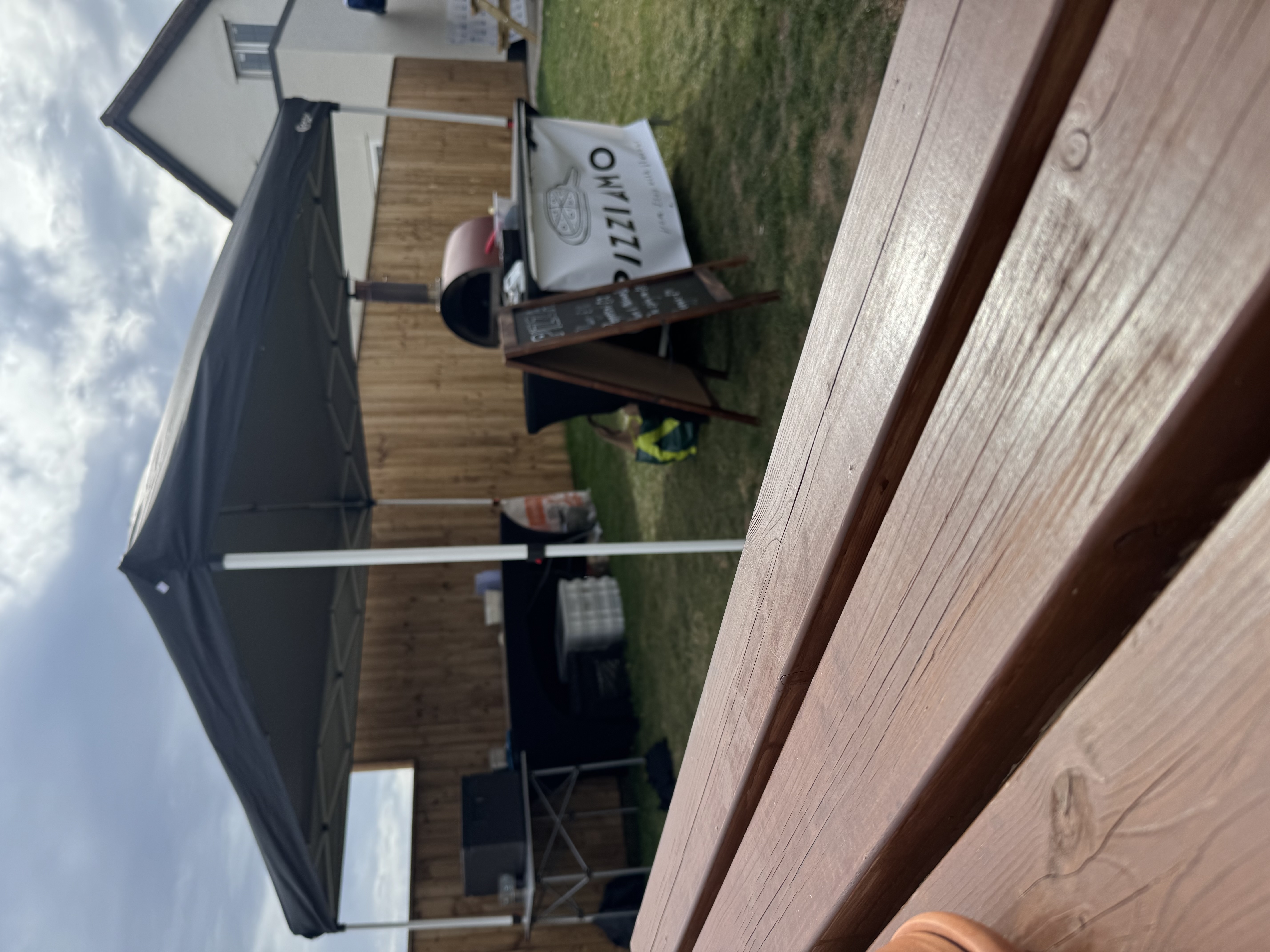 Black canopy tent shelters a Pizziamo catering pizza oven and chalkboard menu on a lawn, with warm wood fencing and bright daylight.