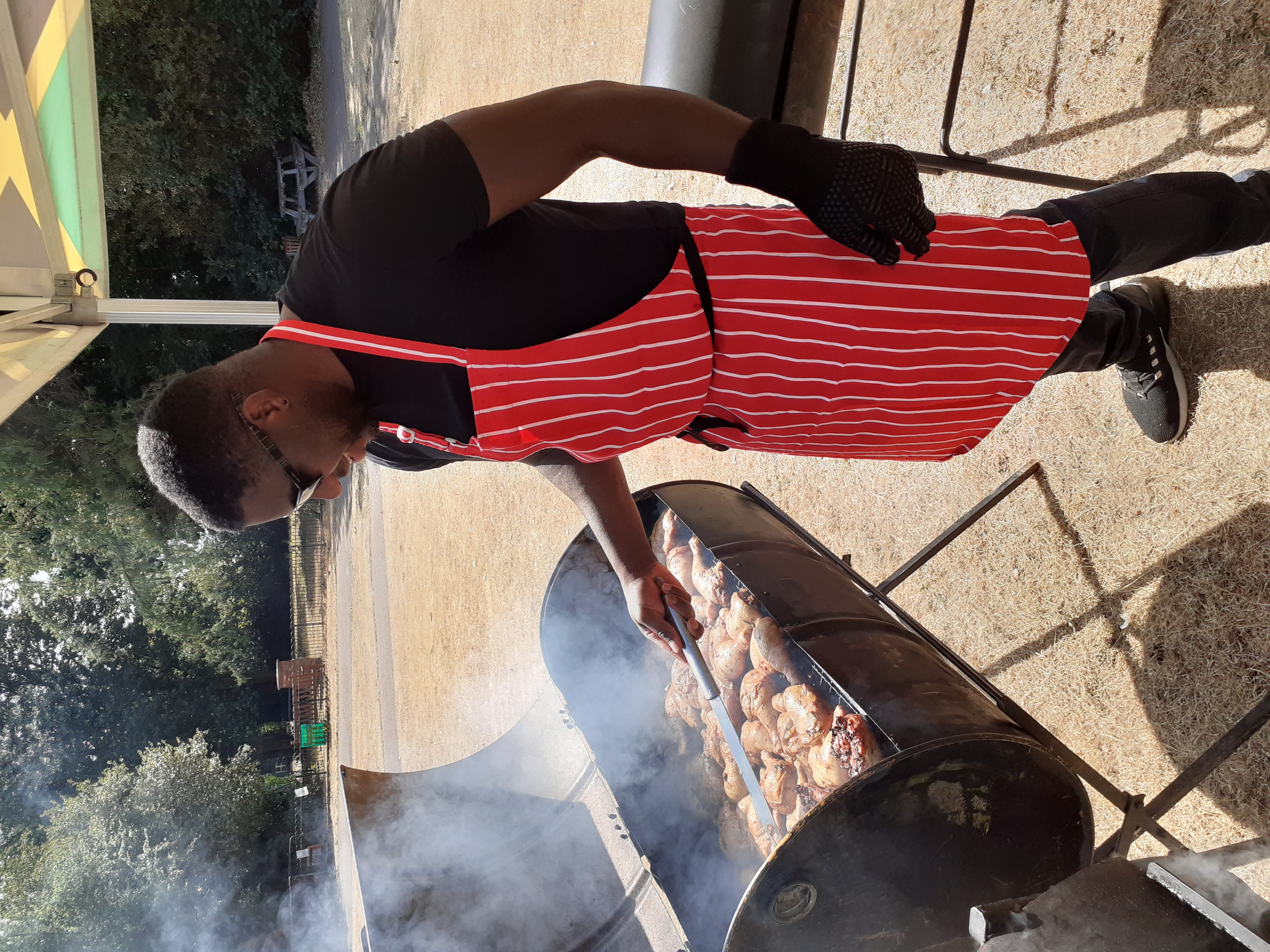 A chef in a red-striped apron grills marinated chicken over a smoky barbecue in bright daylight, showcasing expert catering skills.