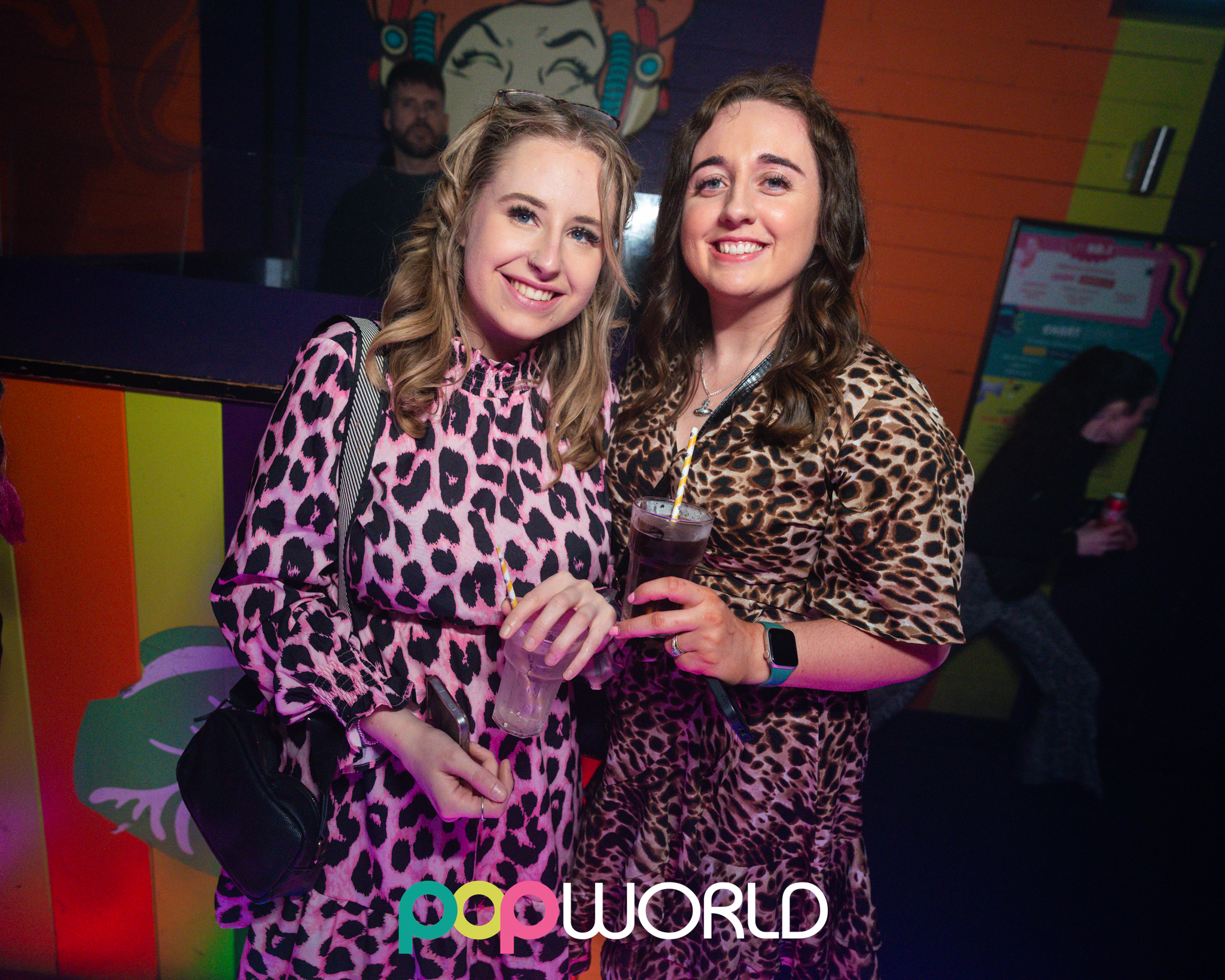 Two smiling women in leopard-print dresses pose at a vibrant nightclub backdrop, holding cocktails under pink-purple lighting at a popworld event.