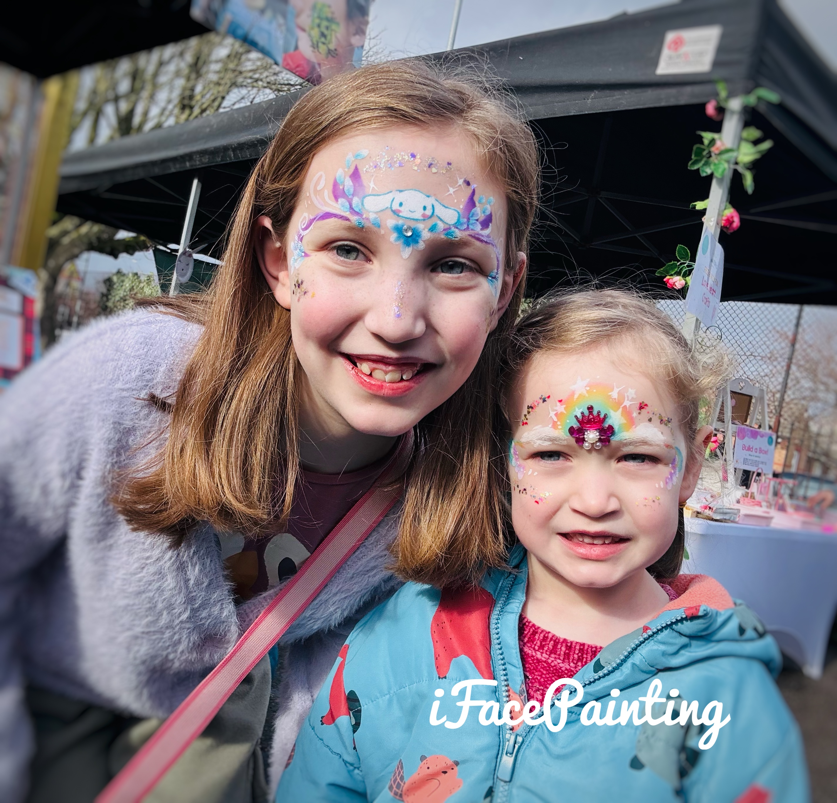 Two girls smile under bright daylight, showcasing colorful face paintings with sparkles. A festive atmosphere surrounds them, hinting at joyful services.