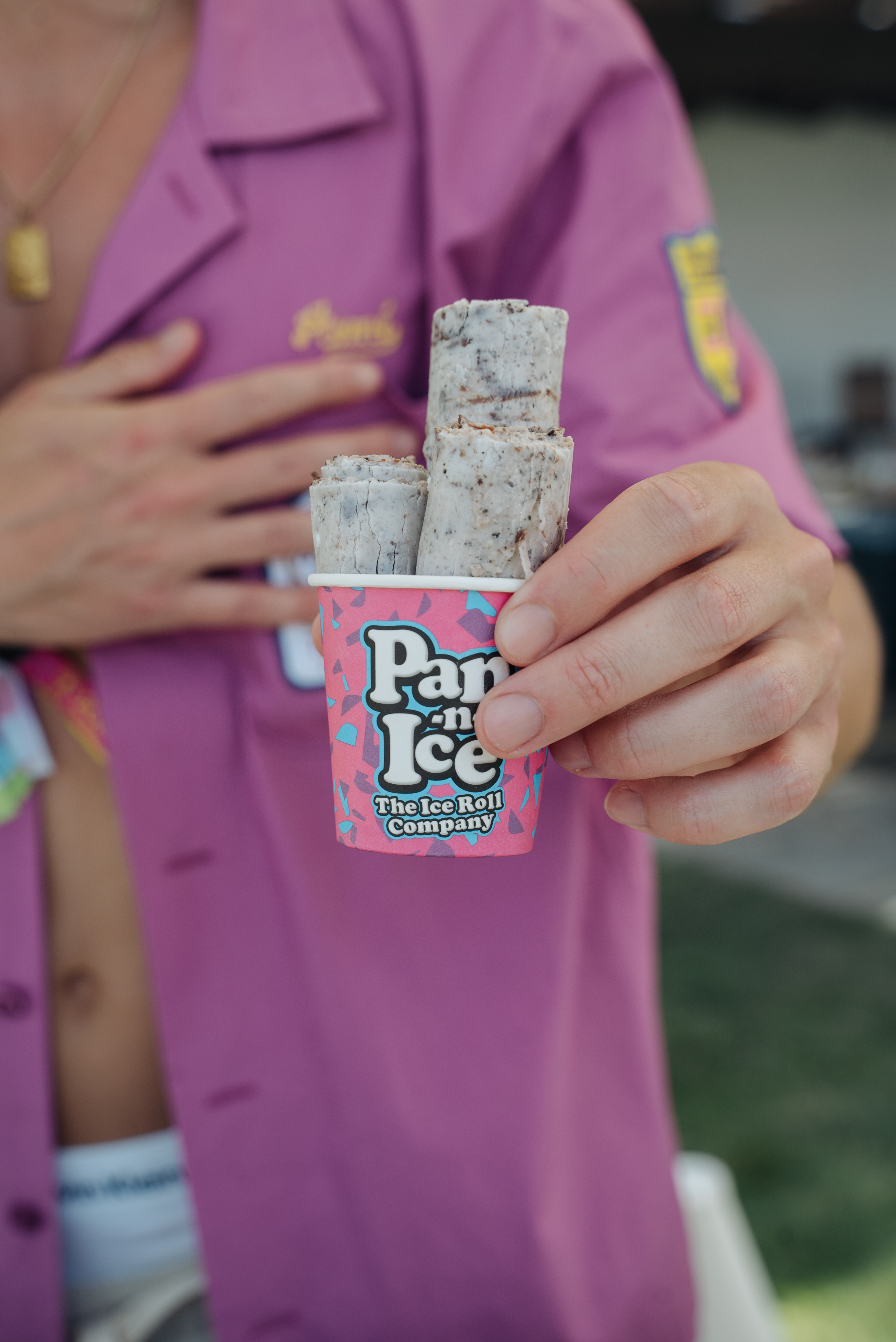 A hand holds a colorful cup of rolled ice cream, featuring creamy, textured rolls with a playful pink design, conveying a fun, vibrant atmosphere.