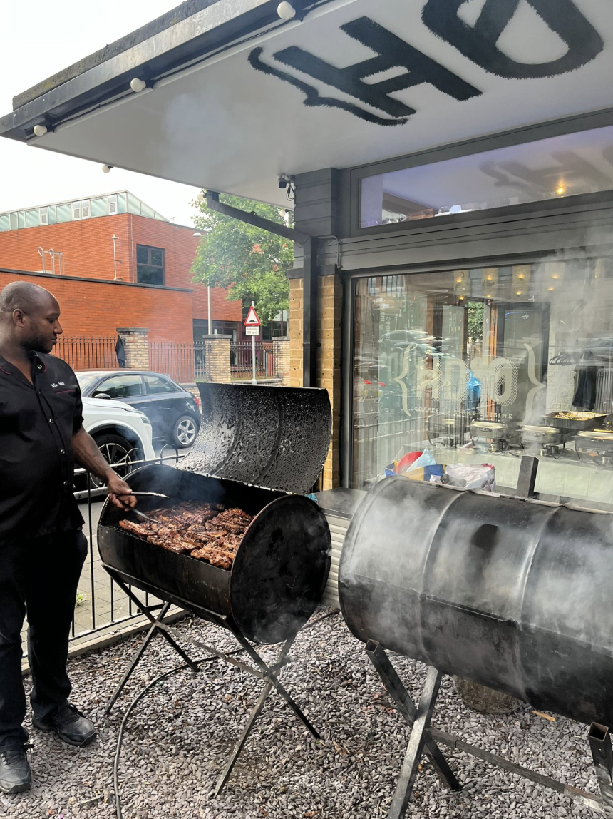 A chef in a black apron tends to smoky, marinated meats on a large charcoal grill, set against a modern, urban backdrop with warm lighting.