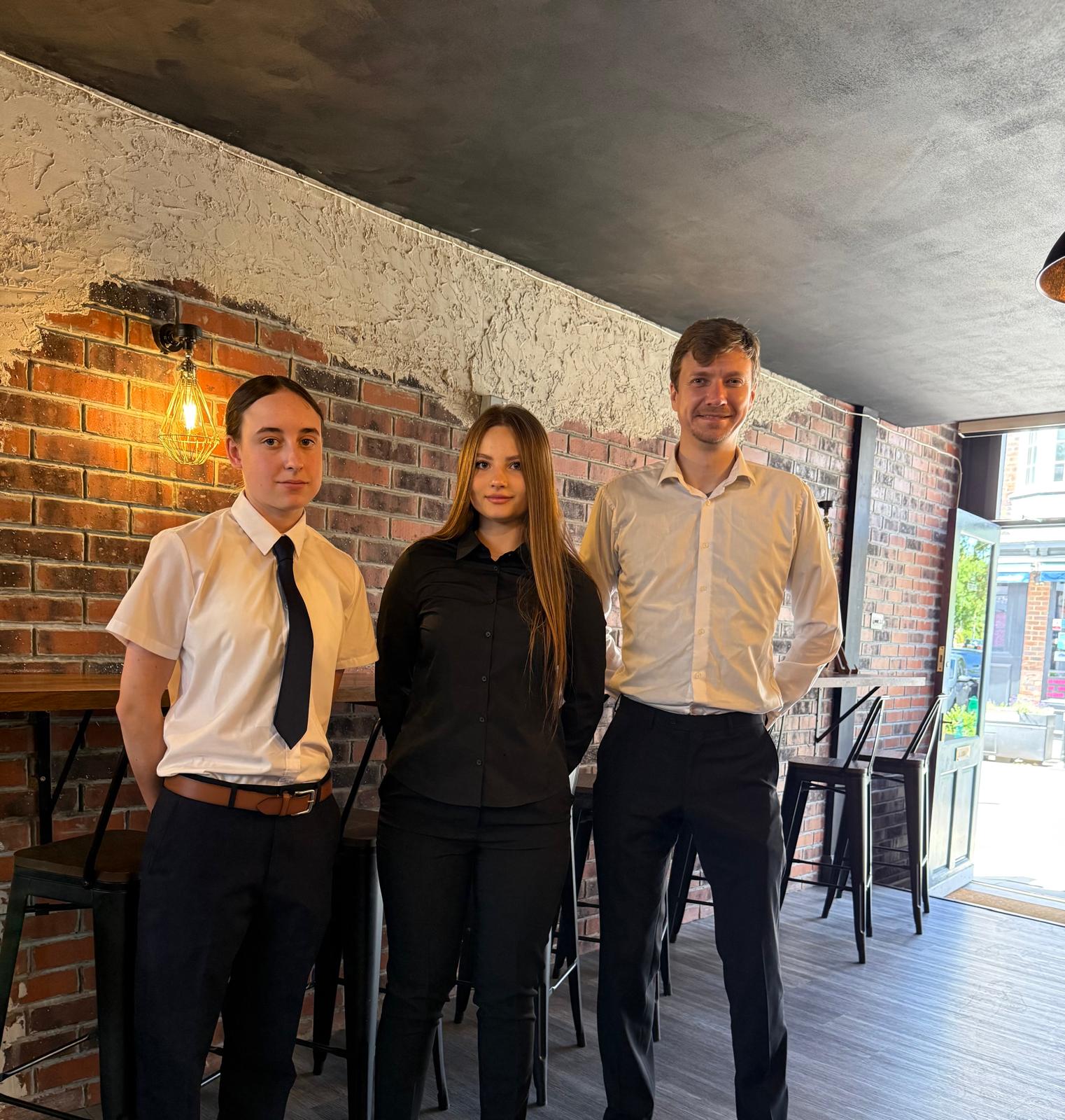 Three uniformed catering staff stand in a cozy brick café, under warm pendant light, beside high tables and polished bar stools.