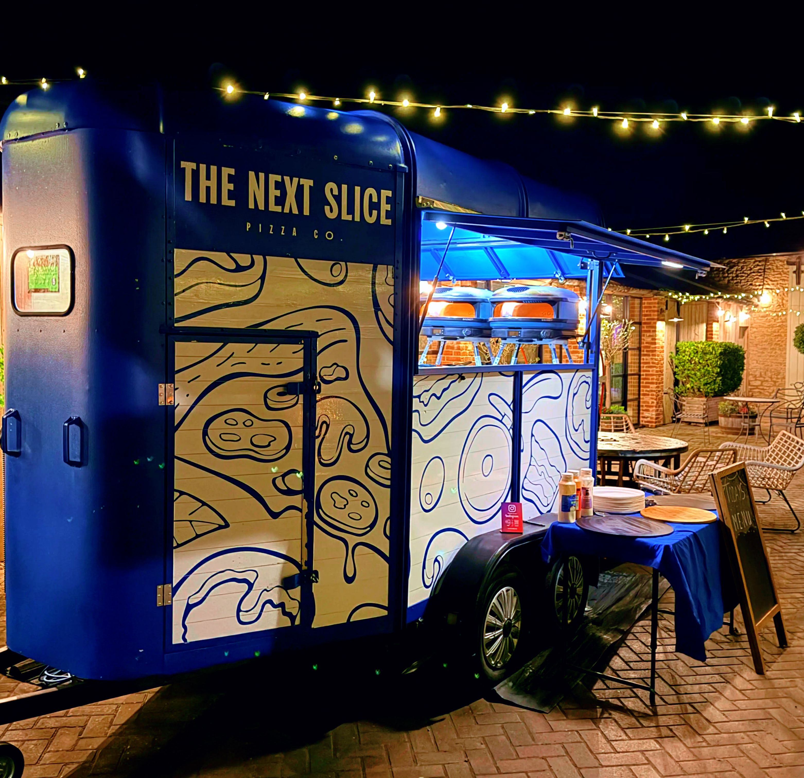A vibrant food truck, "The Next Slice," features a blue façade with playful pizza artwork, illuminated by festive string lights, creating a cozy atmosphere for late-night catering.