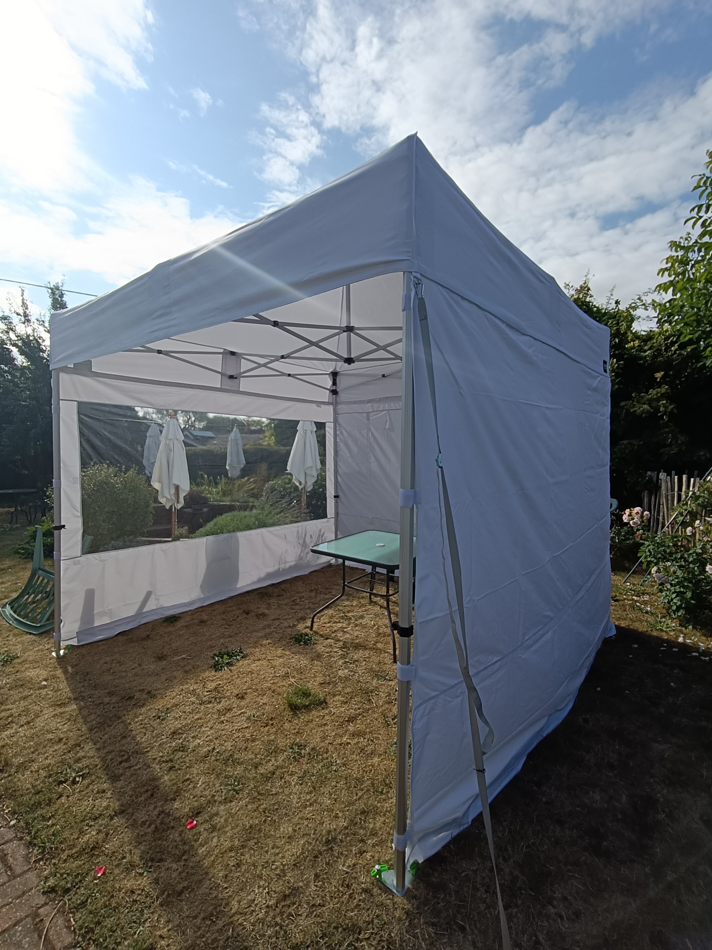 White event marquee tent with clear sidewalls, folded patio umbrellas and a glass-topped table, set in bright daylight on a lawn.