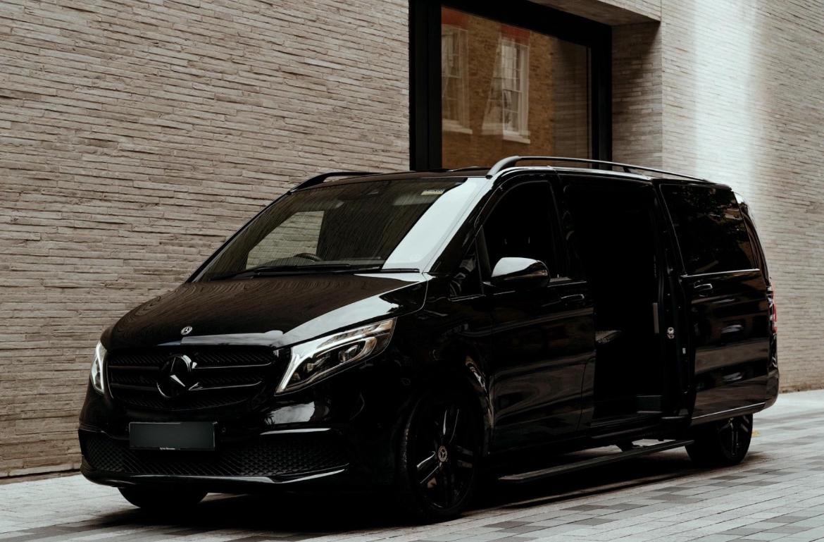 Glossy black Mercedes chauffeur van parked by a beige brick wall, side door open, sleek luxury transport under soft daylight.