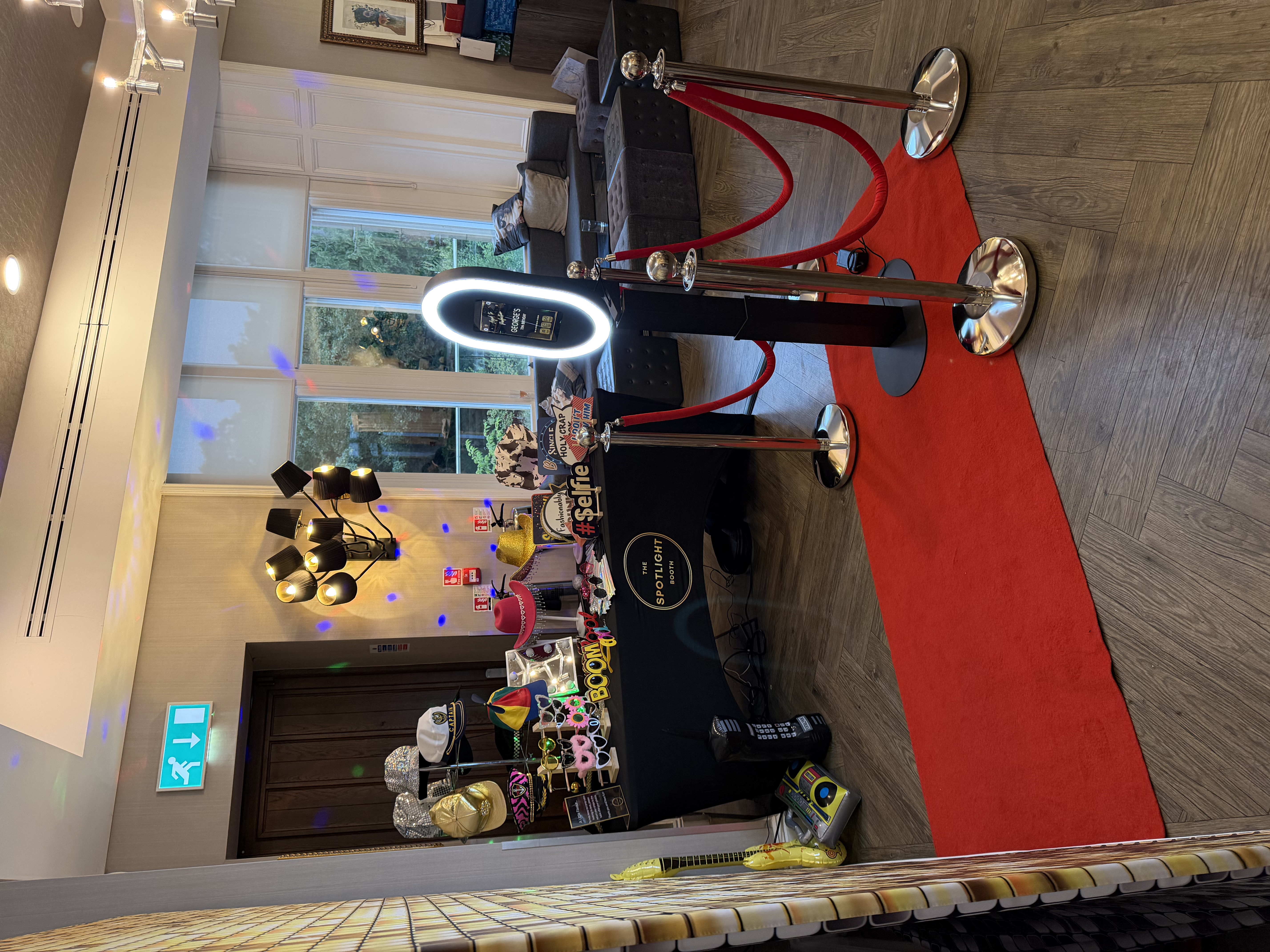 Bright event hall with a red carpet, ring-light photo booth, props table, velvet stools, and polished stanchions in warm daylight.