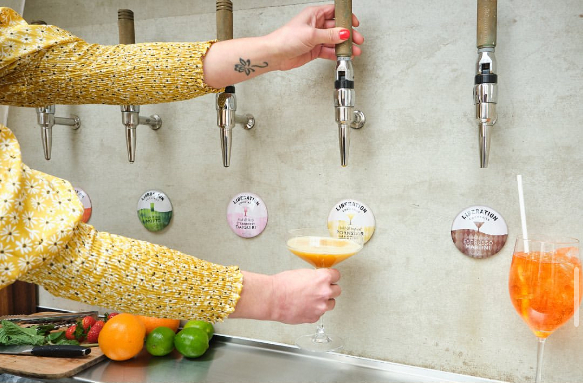 A vibrant yellow floral sleeve holds a cocktail glass by a sleek, stainless-steel bar with colorful drink options, evoking lively service vibes.