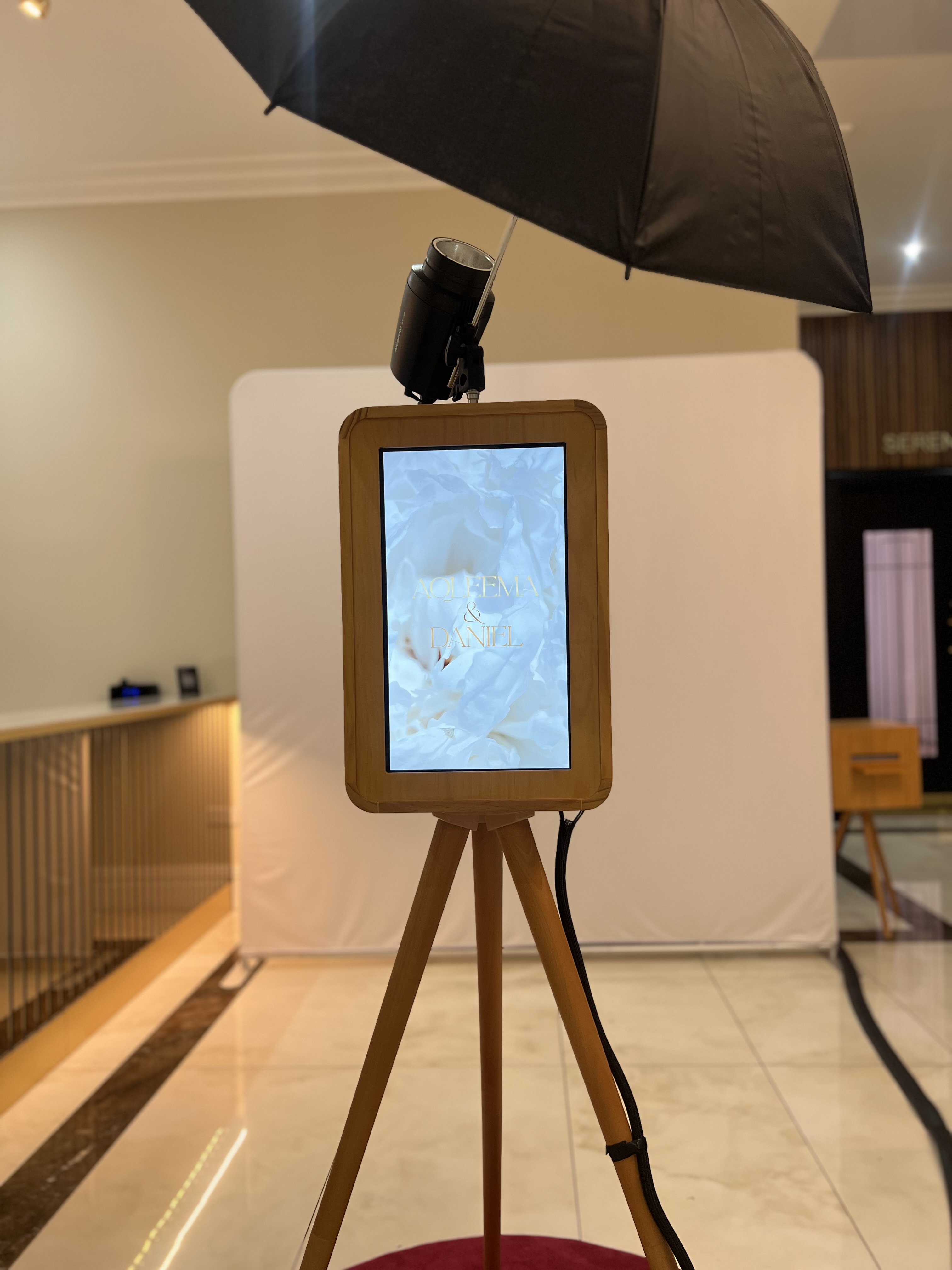 A wooden photo booth stands on a red carpet, draped under soft lighting with a black umbrella. The backdrop is an elegant white screen.