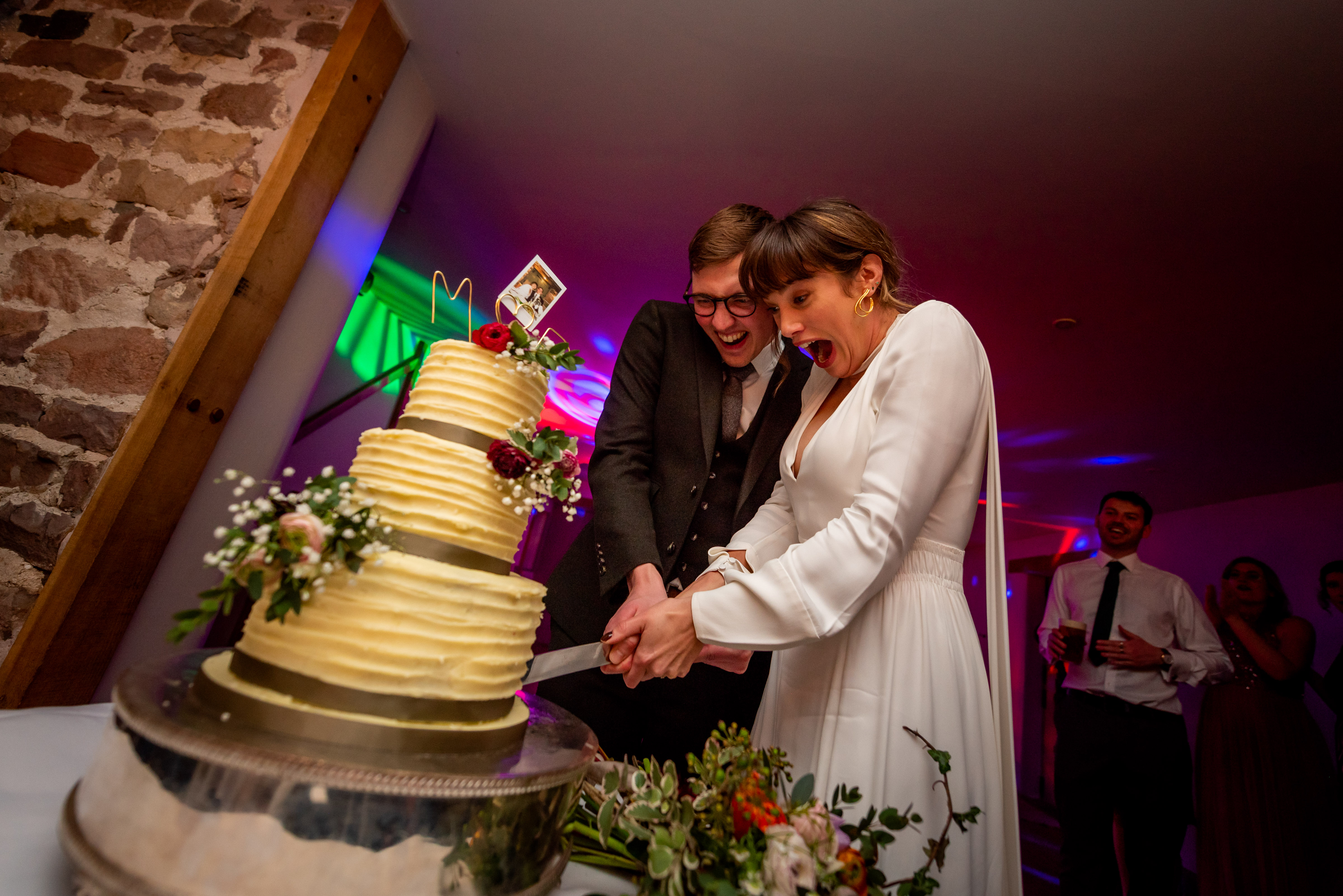 A couple joyfully cuts a tall, frosted, three-tier wedding cake adorned with delicate flowers under vibrant party lights, celebrating love.