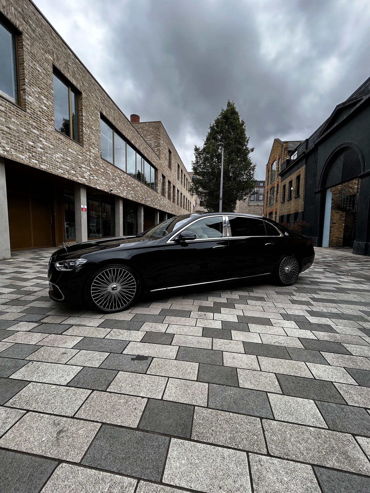 A sleek black luxury sedan is parked on stylish gray and white patterned cobblestone, surrounded by modern brick buildings under overcast skies.