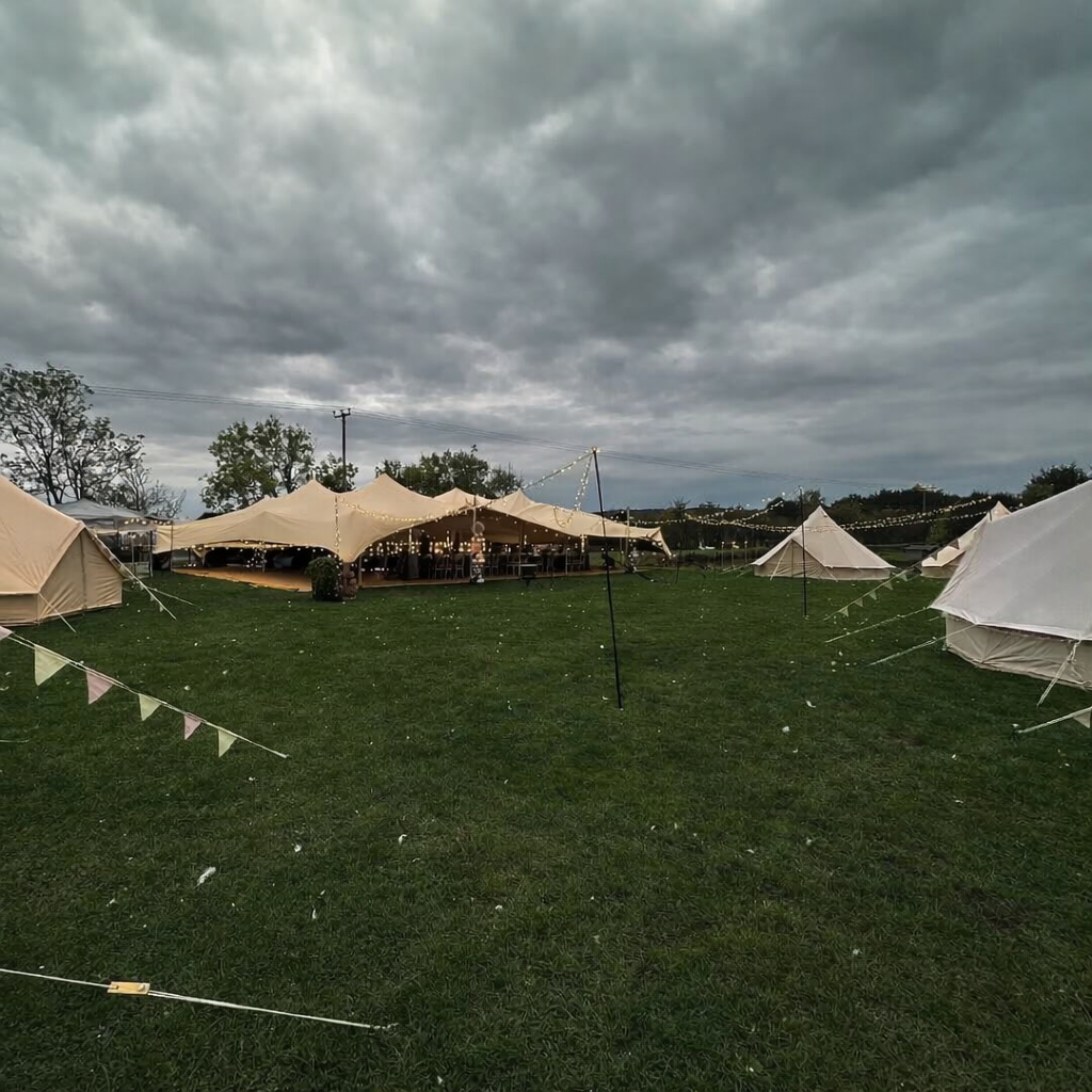 A spacious outdoor event site features multiple beige tents and a central decorated dining area with warm string lights, set against a moody sky.