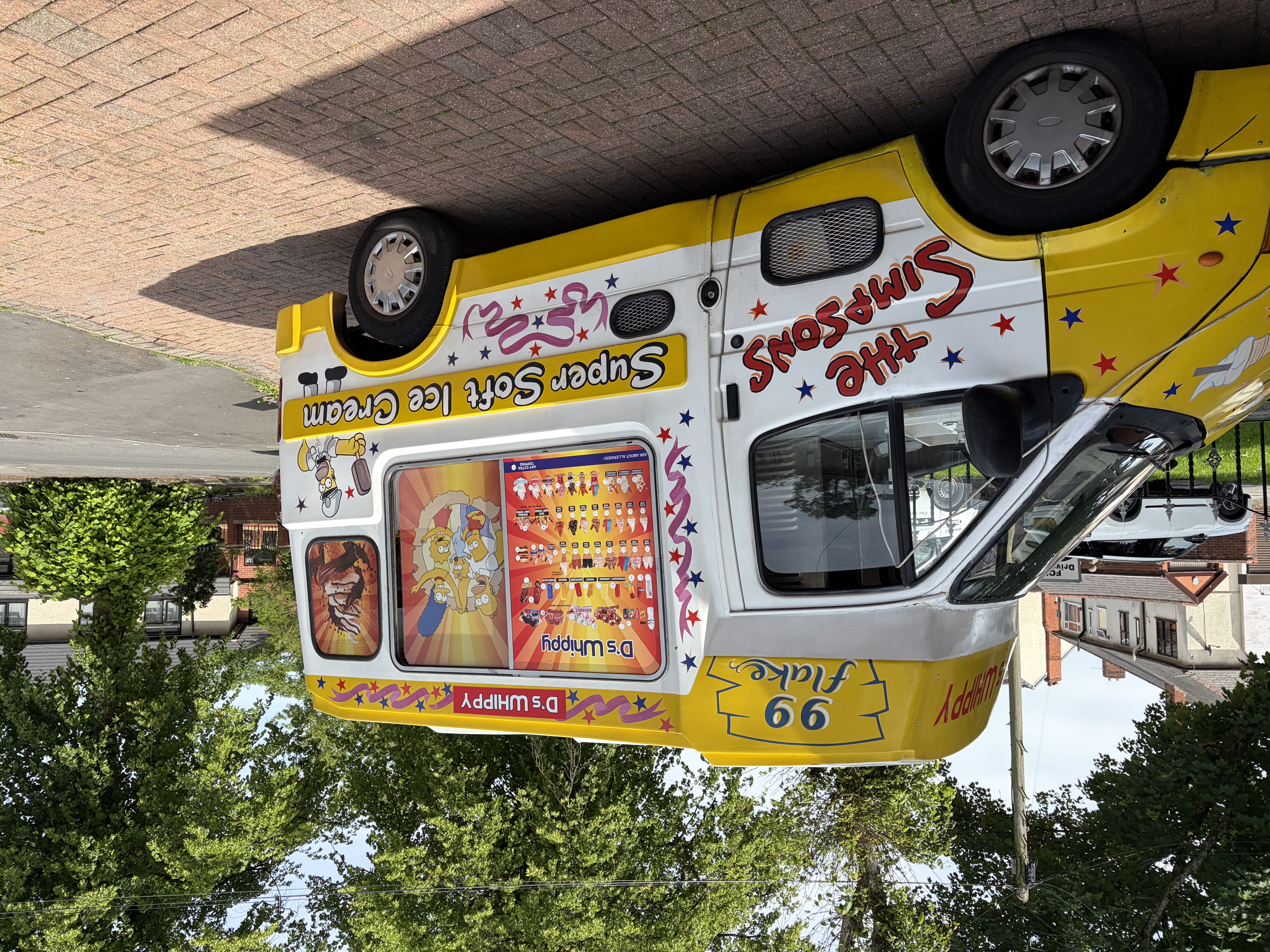 Bright daylight reveals a yellow-and-white Simpsons soft ice cream catering van, parked neatly on a brick drive amid leafy trees.
