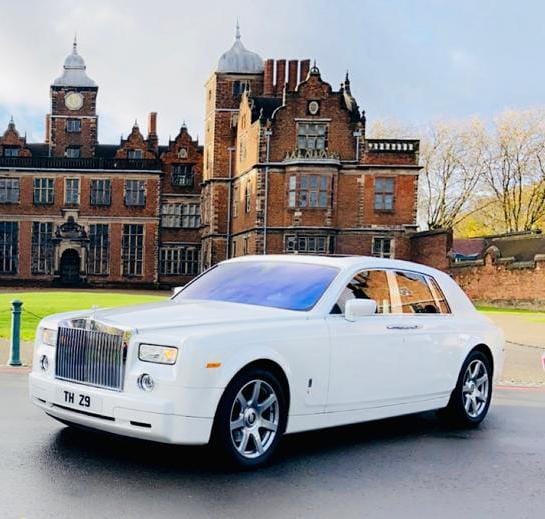 A sleek white Rolls-Royce shines against a grand vintage brick estate, with elegant towers and intricate architecture, under soft daylight.