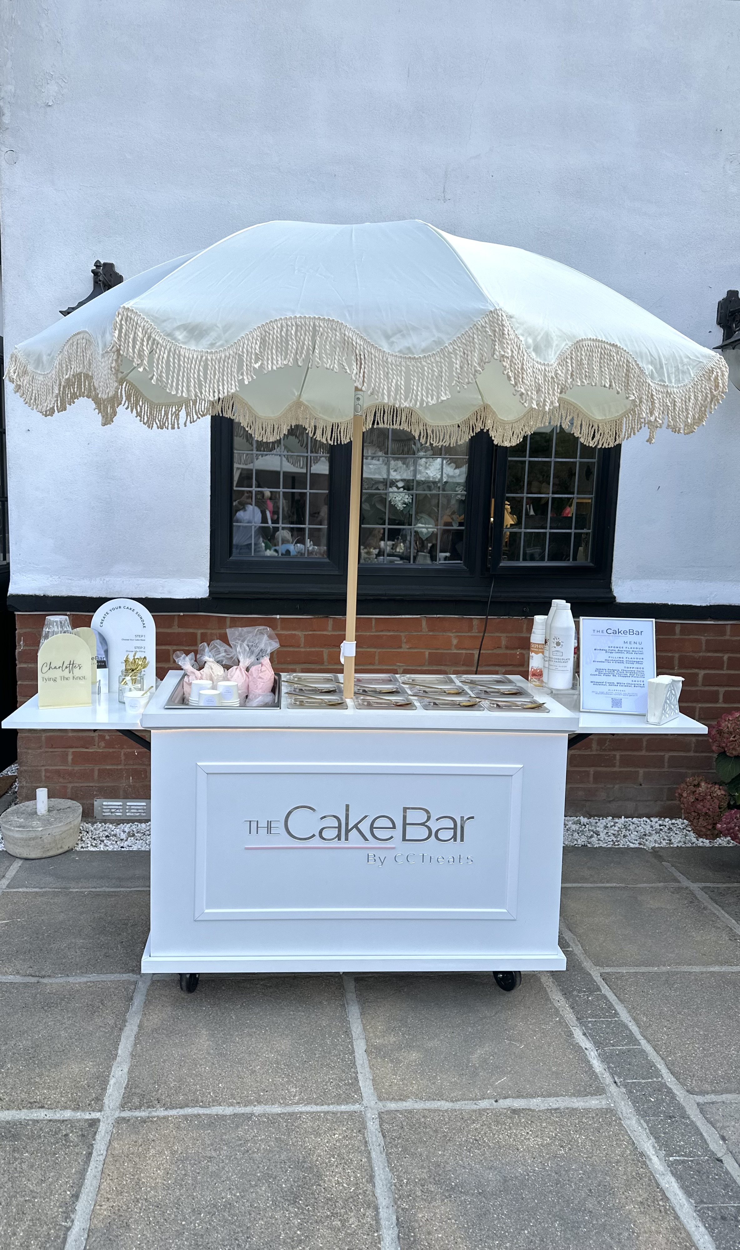 A pristine white cake bar featuring a fringed umbrella sits on a stone patio, showcasing delicately arranged sweets under soft daylight.