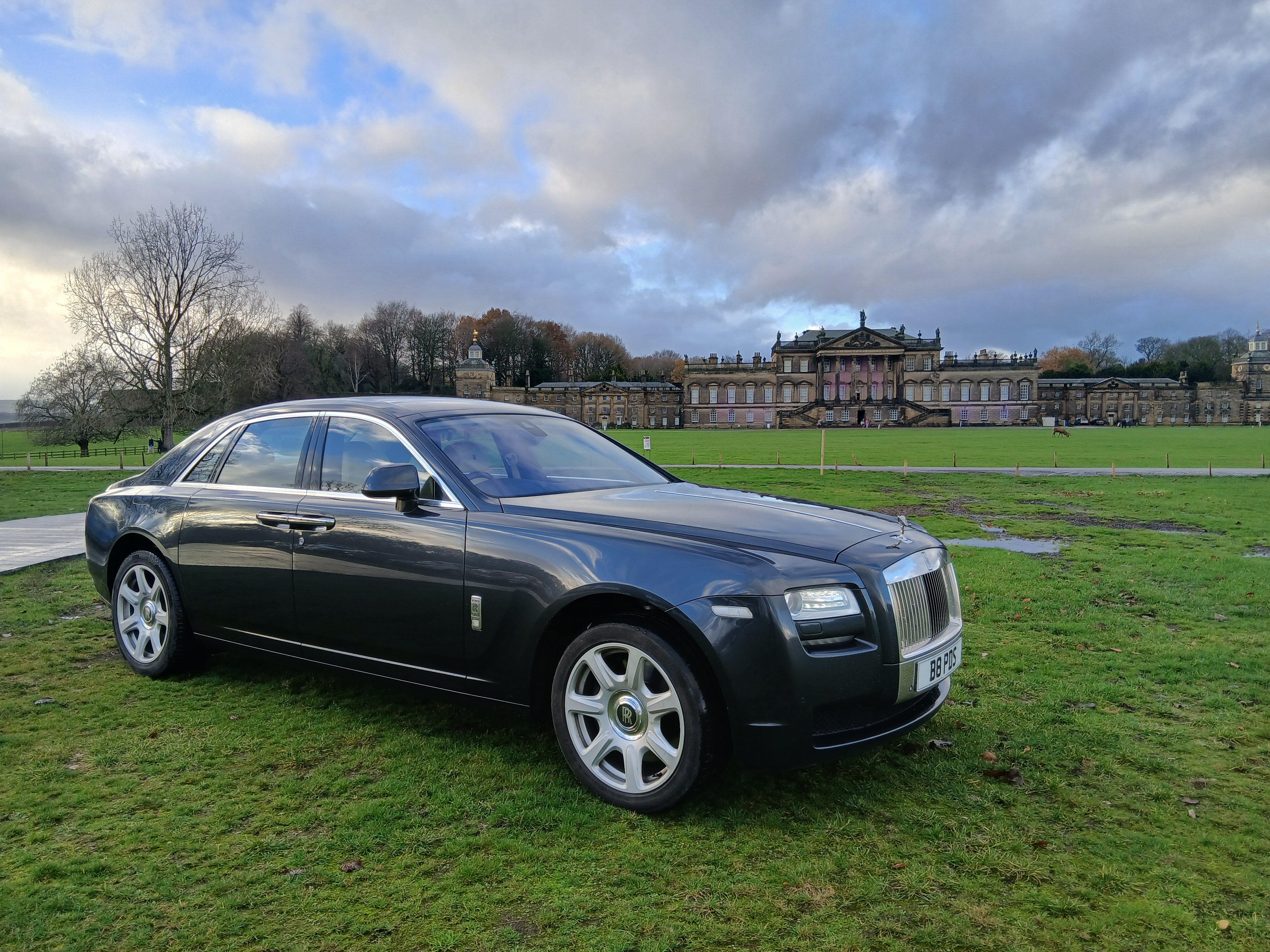 Black Rolls-Royce sedan on wet grass before a grand manor, polished chrome gleaming under cloudy daylight, luxury chauffeur-service feel.